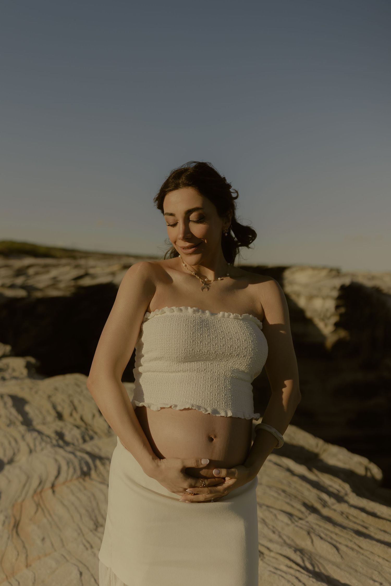 Pregnant woman in white crop top and skirt cradling her baby bump outdoors on rocky terrain at golden hour.