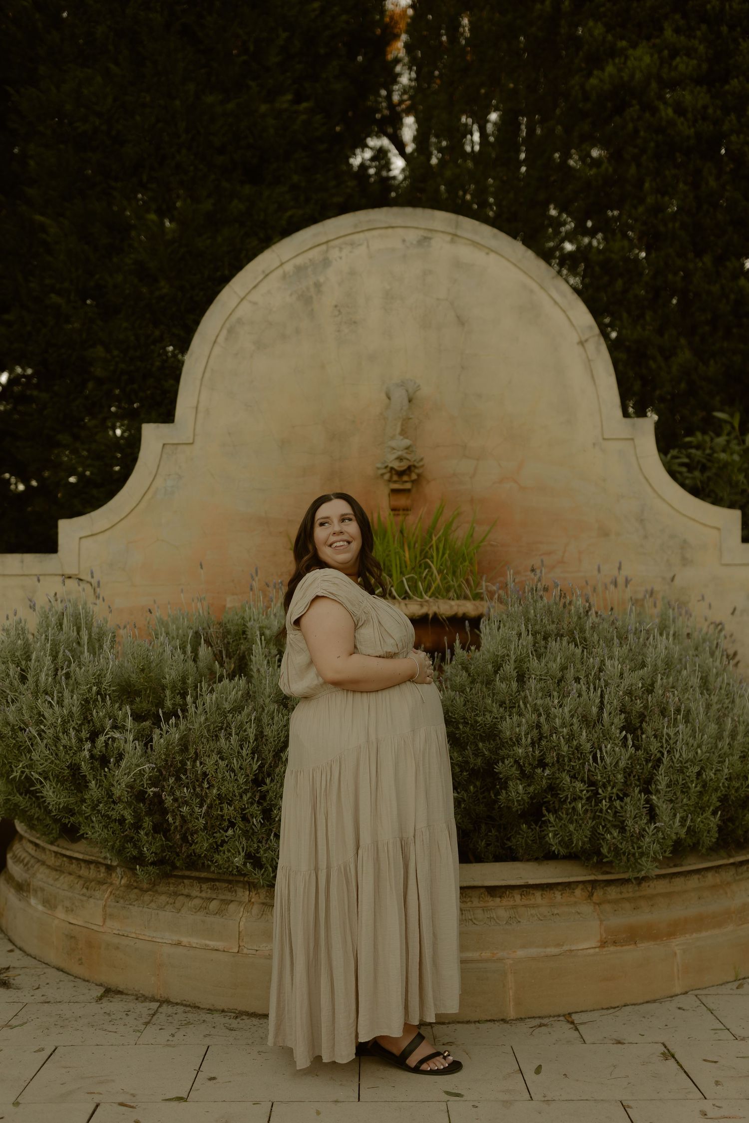 Pregnant woman in flowing cream dress smiles beside an ornate stone fountain surrounded by lavender bushes.