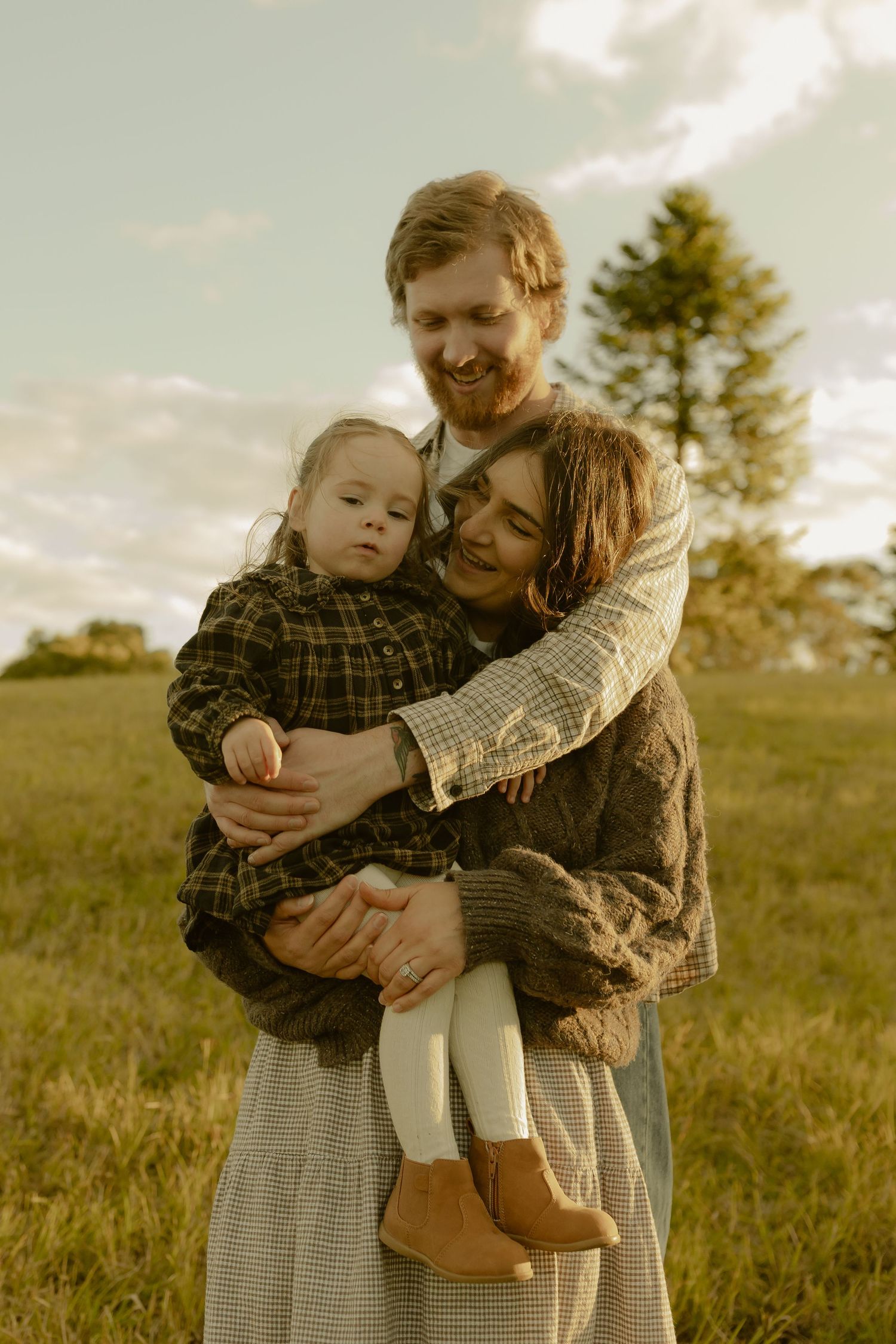 Happy family embracing outdoors, father hugging mother holding toddler in plaid jacket at sunset.