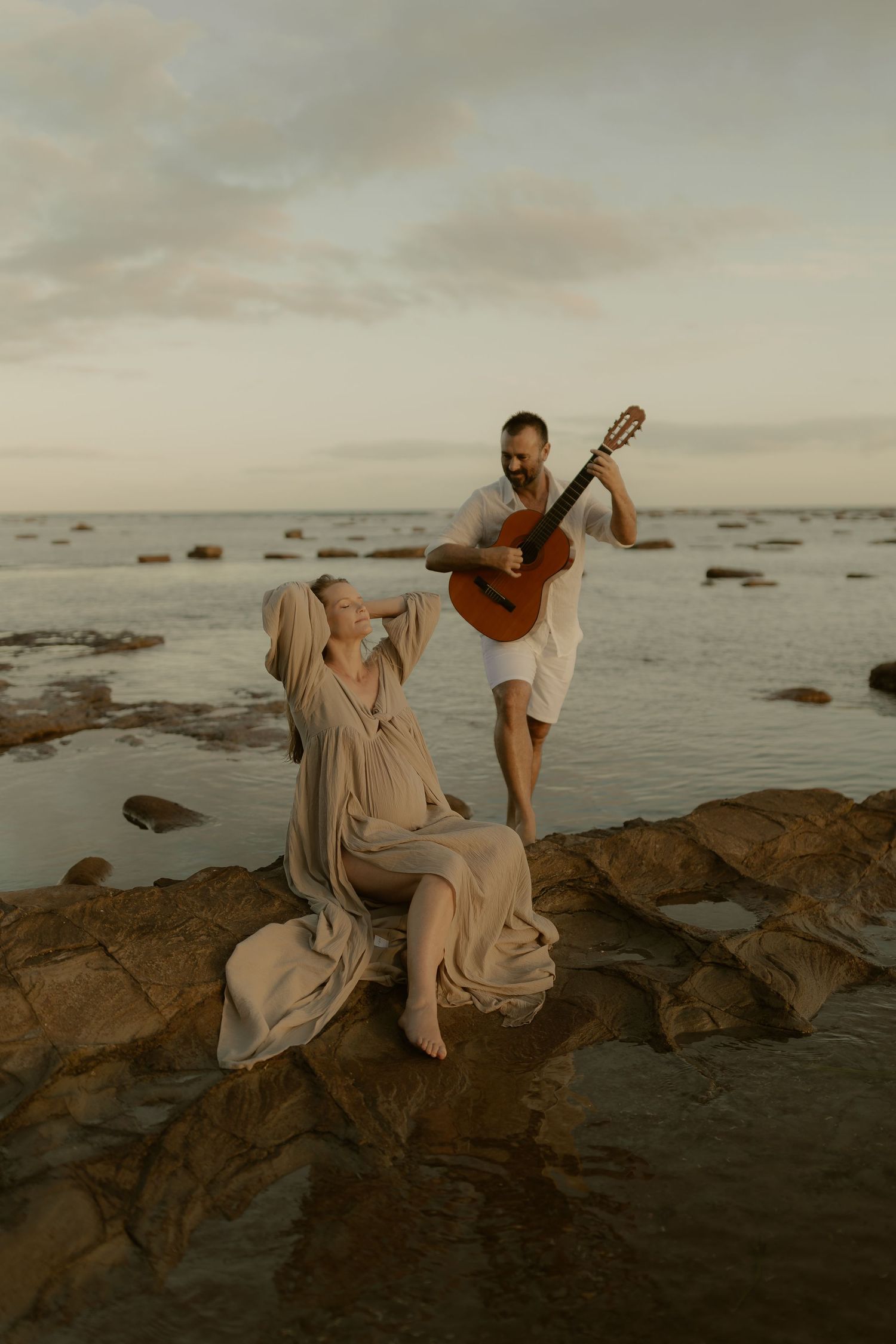 Figure in flowy beige dress poses gracefully with acoustic guitar on rocky coastal landscape at sunset.