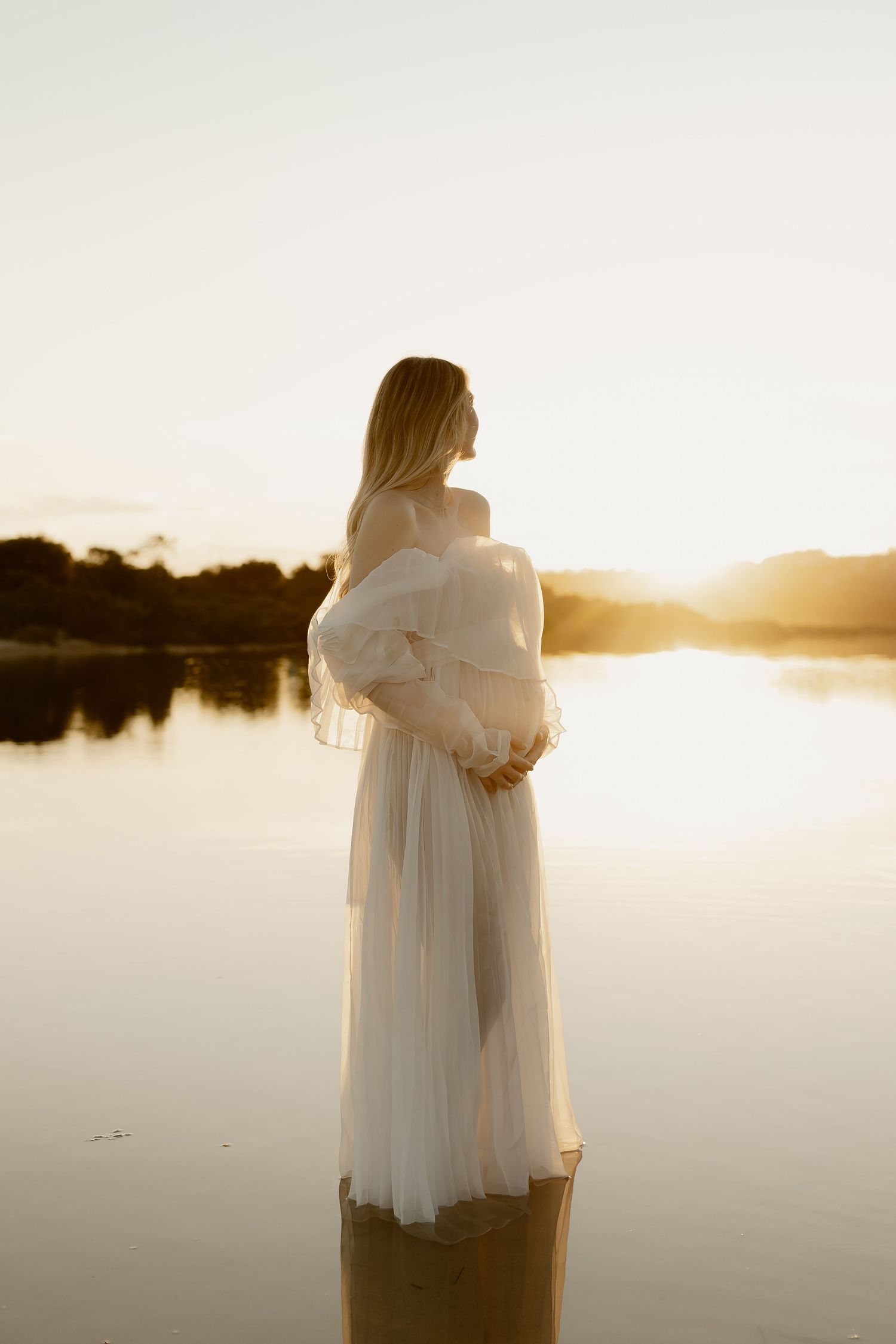 A silhouetted figure in flowing white dress stands in still water during a serene sunset moment.