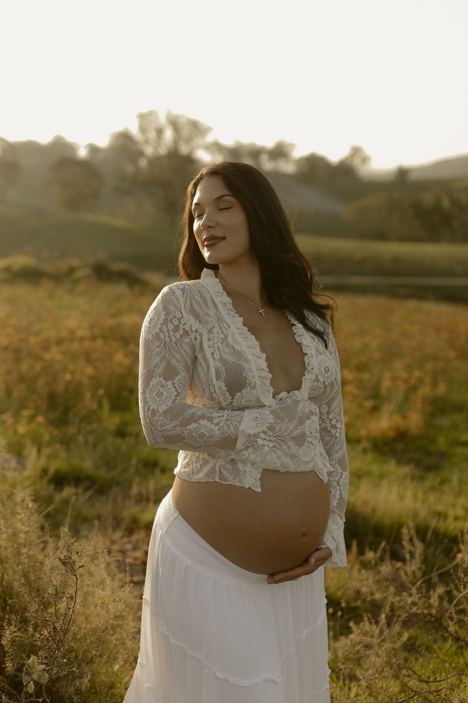 An expectant mother in white lace poses in a sunlit meadow during golden hour.