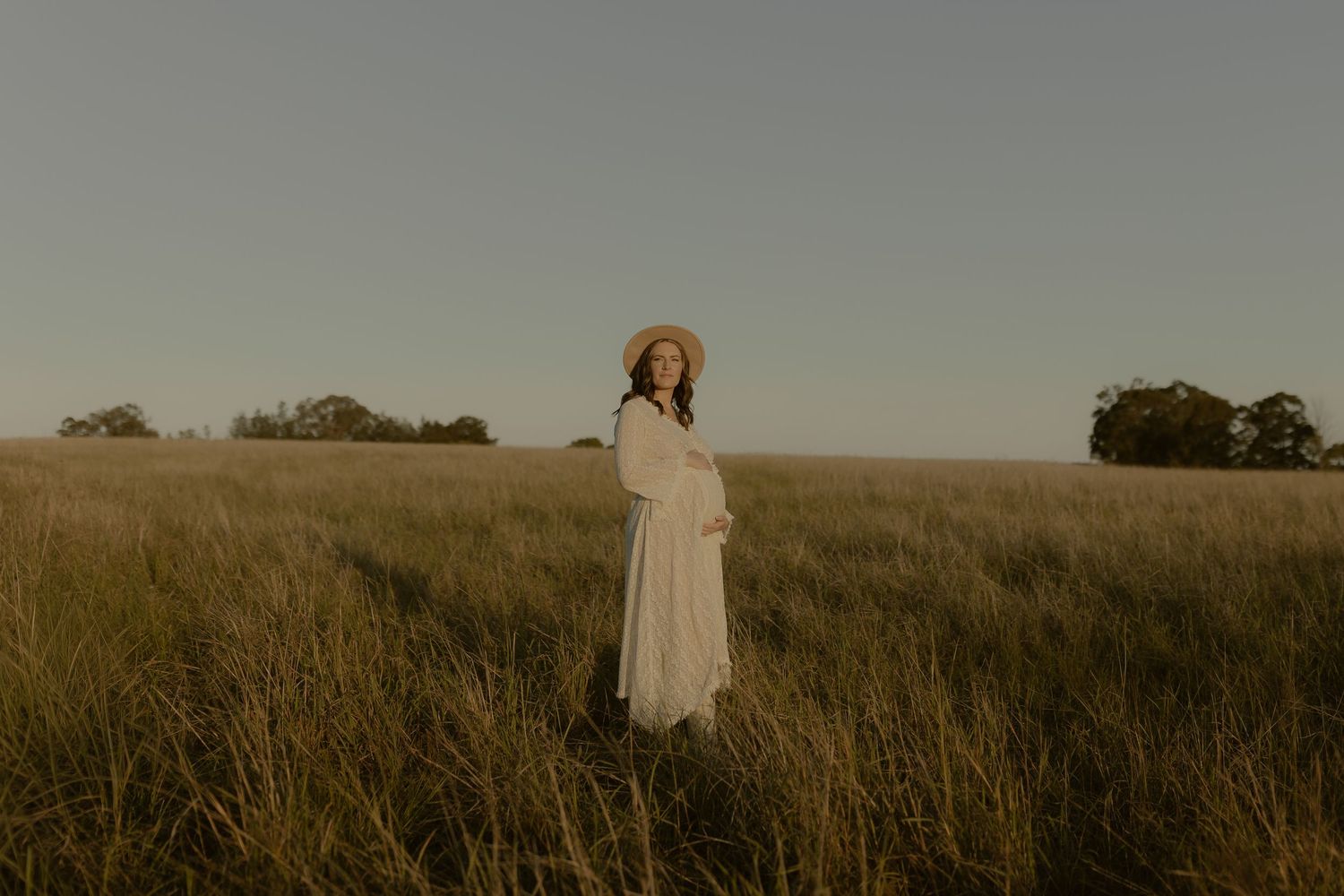 A person in a white dress walks through tall grass in a golden field at sunset.
