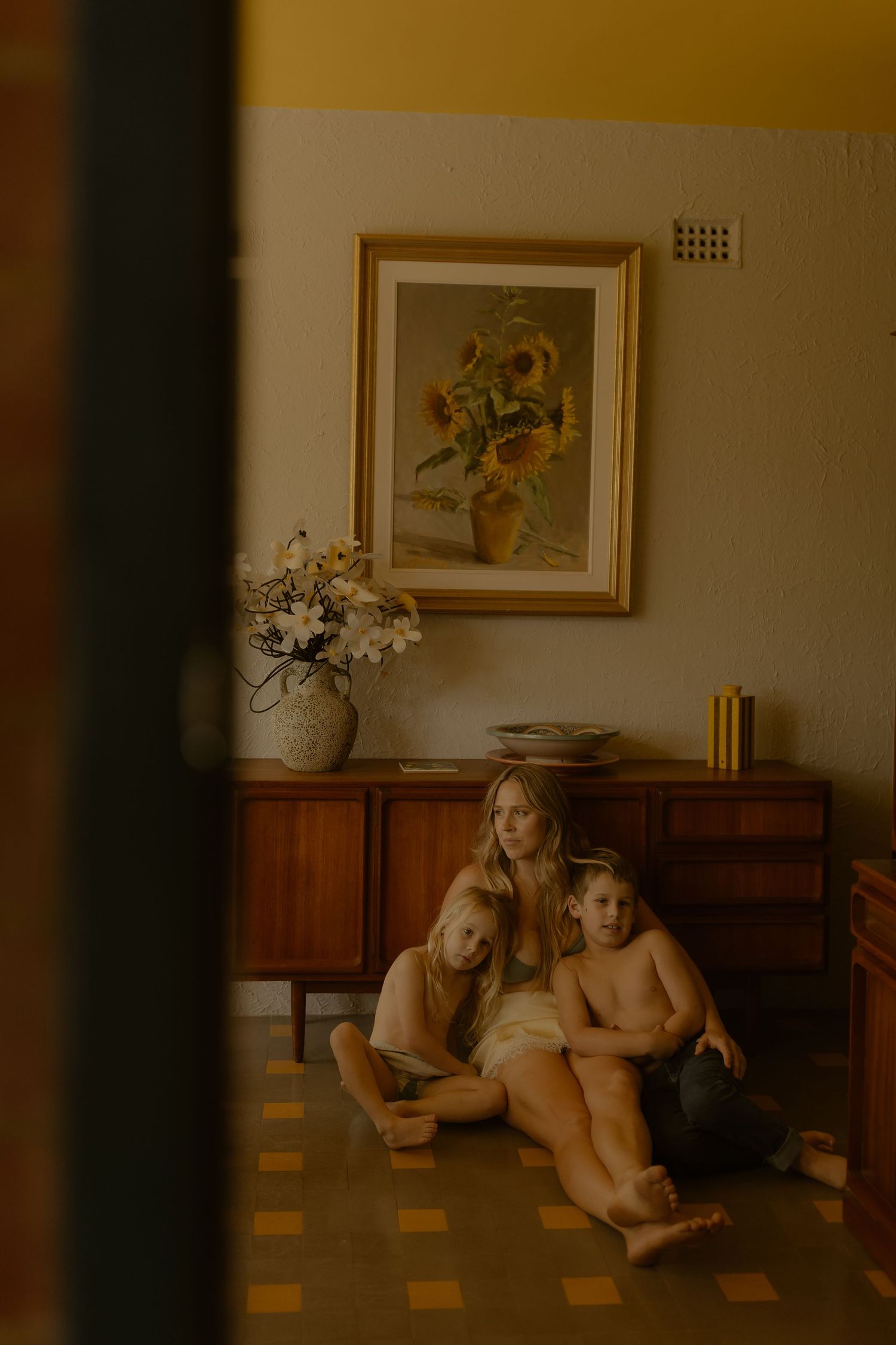 Mother sitting on floor with two children leaning against a wooden sideboard with sunflower painting above.