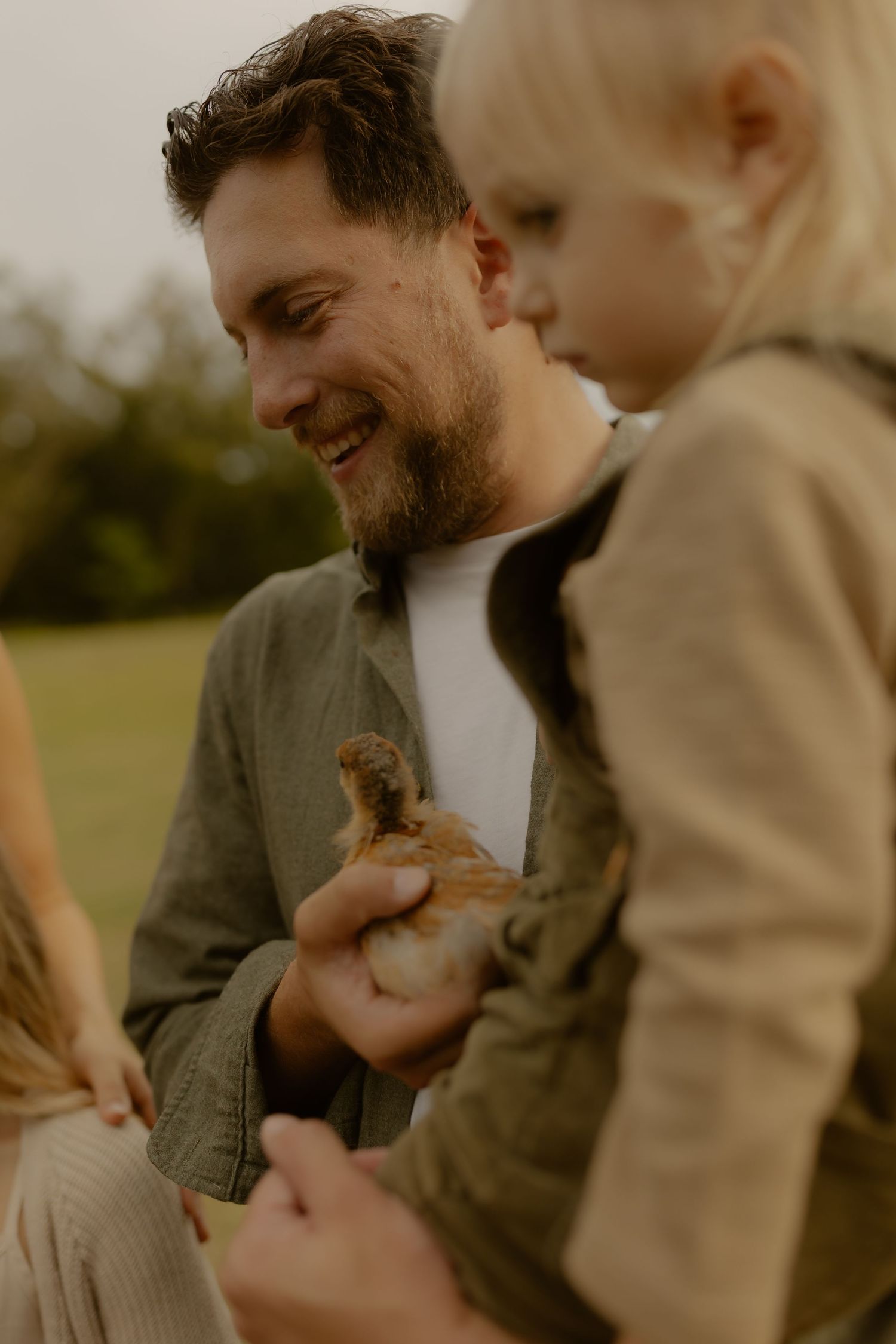 A smiling bearded man holds a baby chick while a young blonde child looks on outdoors.