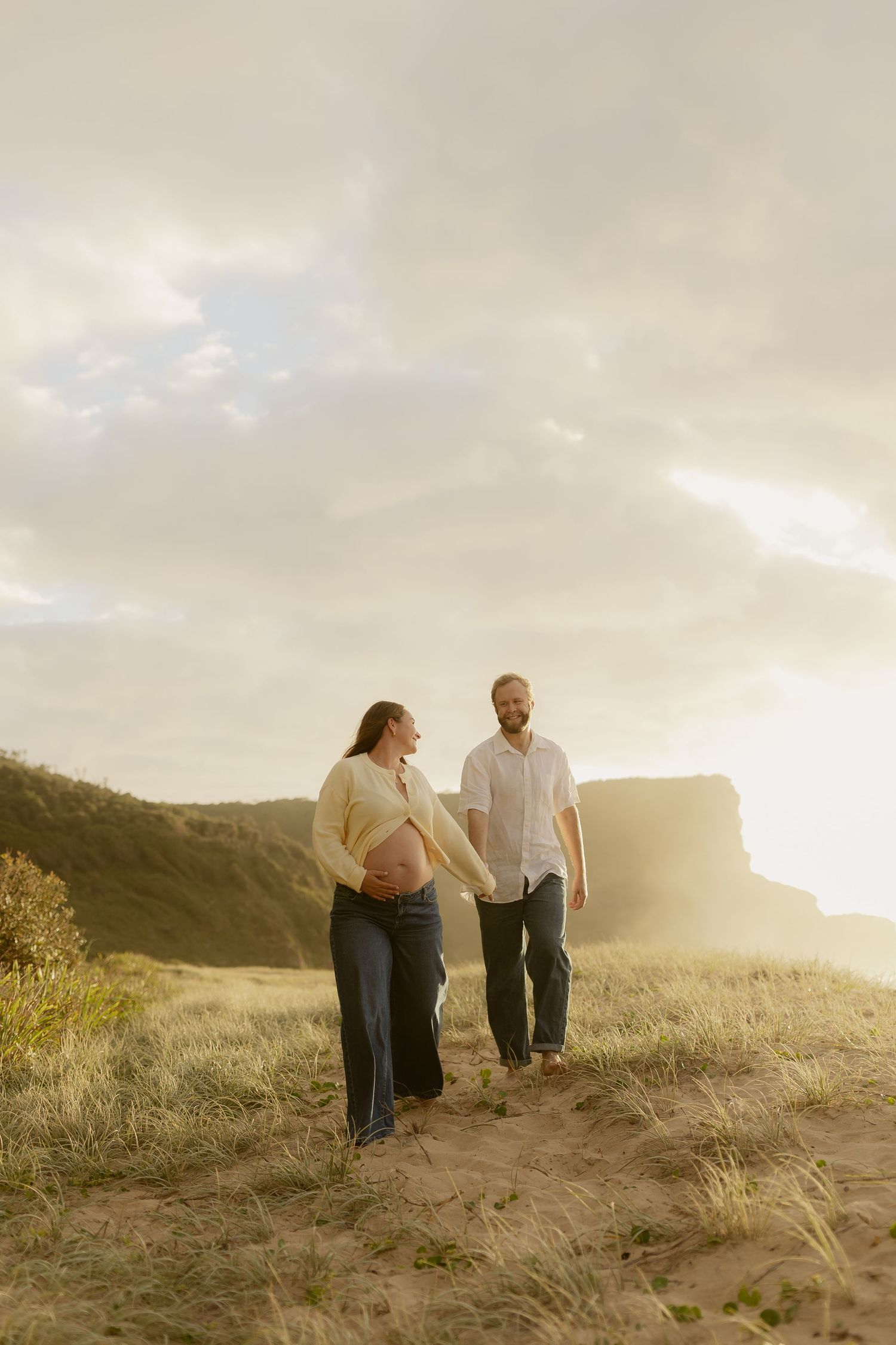 Pregnant couple holding hands walking on sandy coastal dunes at golden hour with cliffs in background.