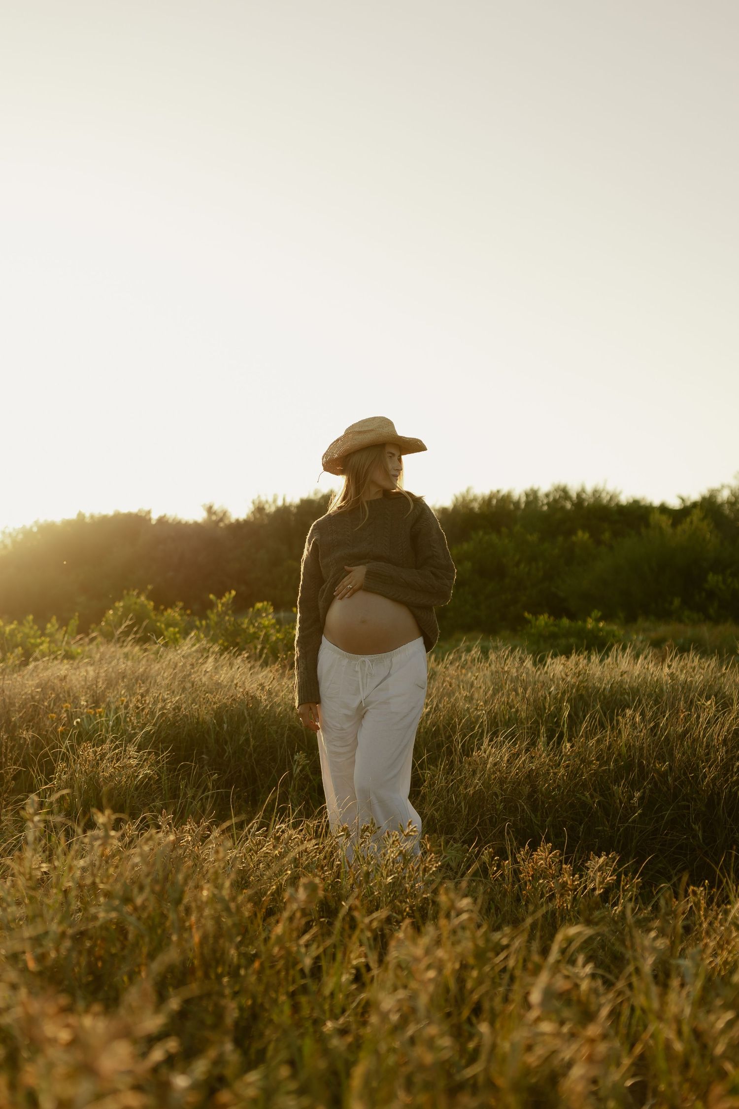 Pregnant woman in white pants and hat standing in golden field at sunset, cradling baby bump.
