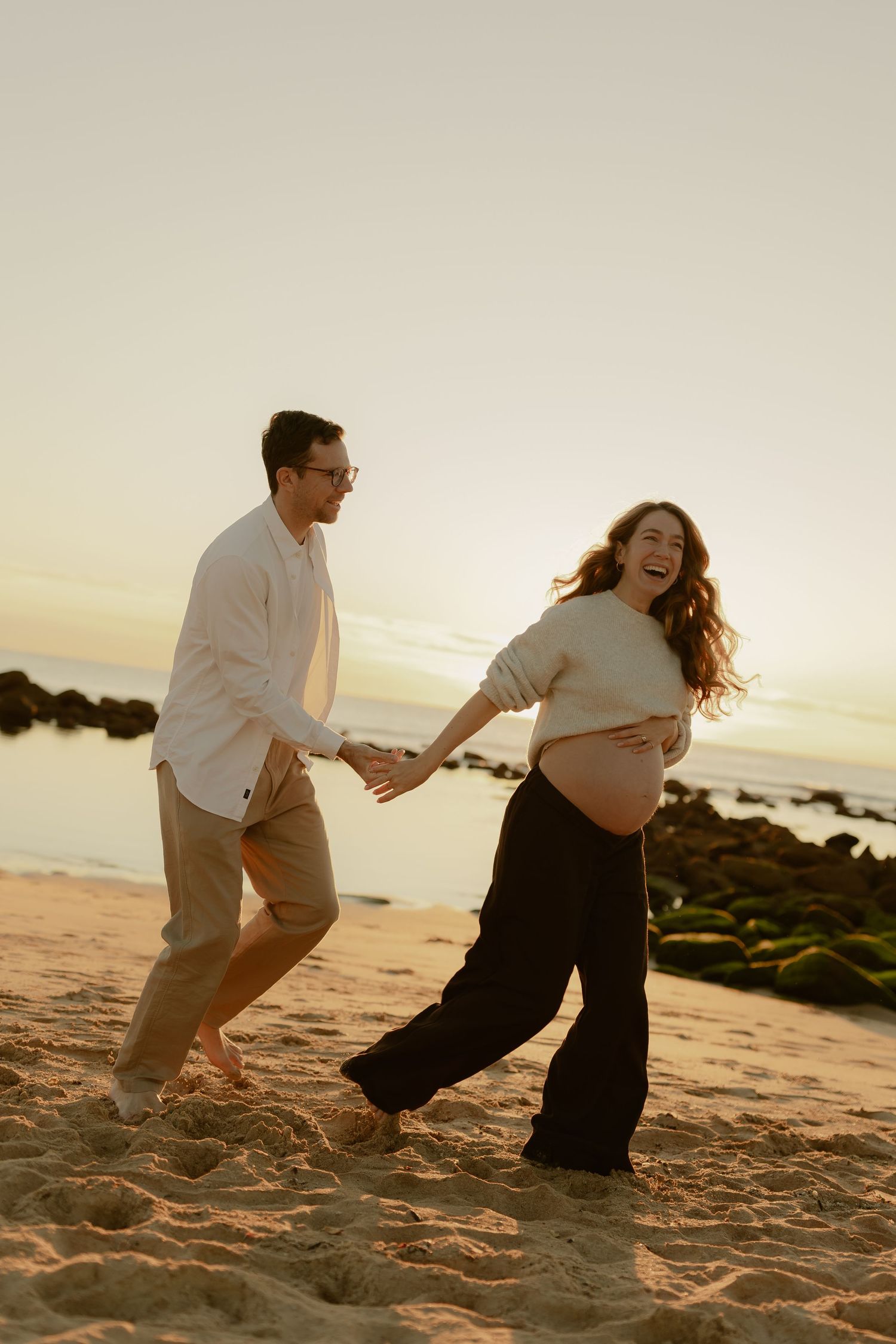 Happy pregnant couple holding hands and laughing on a sandy beach at sunset, maternity photo session.