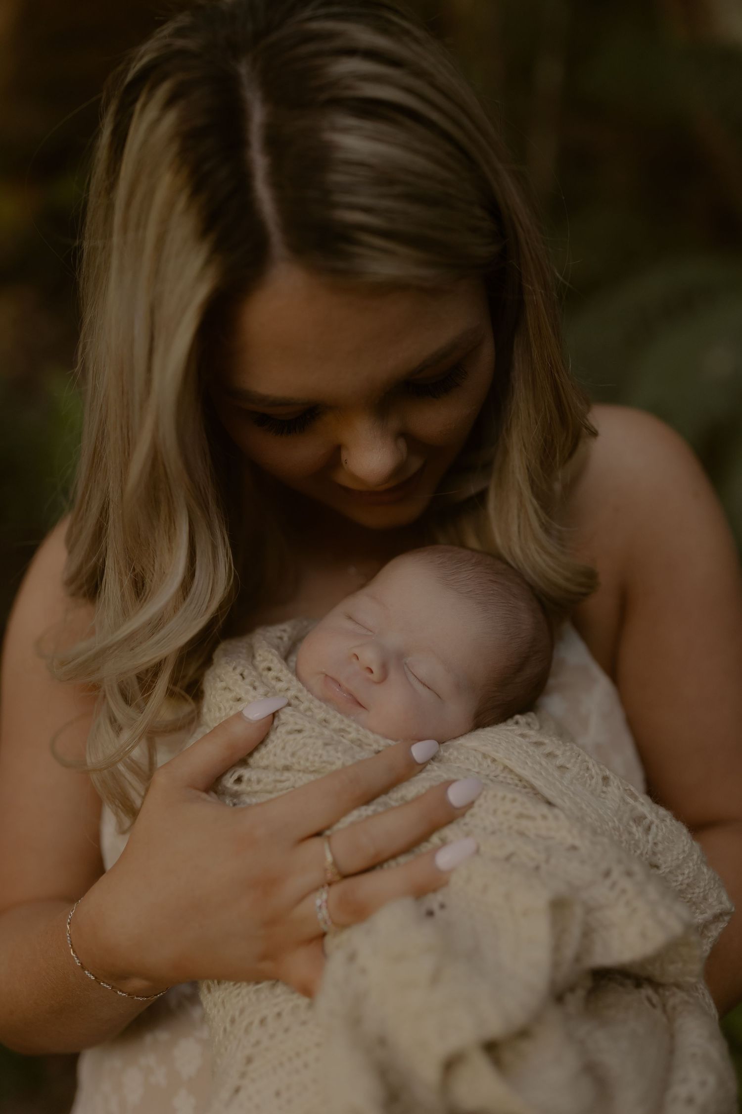 Mother with blonde hair gently cradles sleeping newborn wrapped in cream knit blanket, smiling softly.