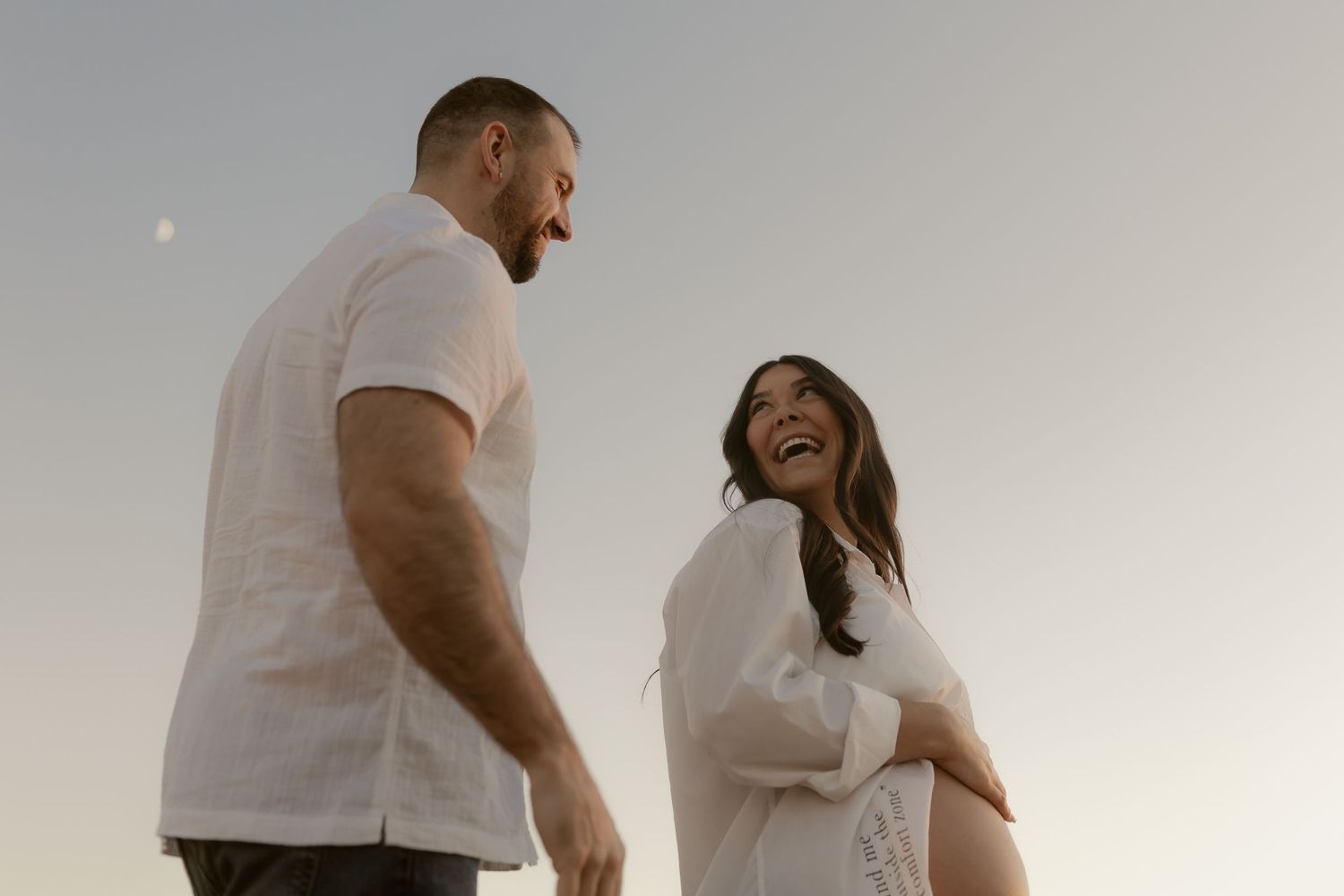 Pregnant couple in white outfits laughing together outdoors at dusk with a crescent moon visible in the sky.