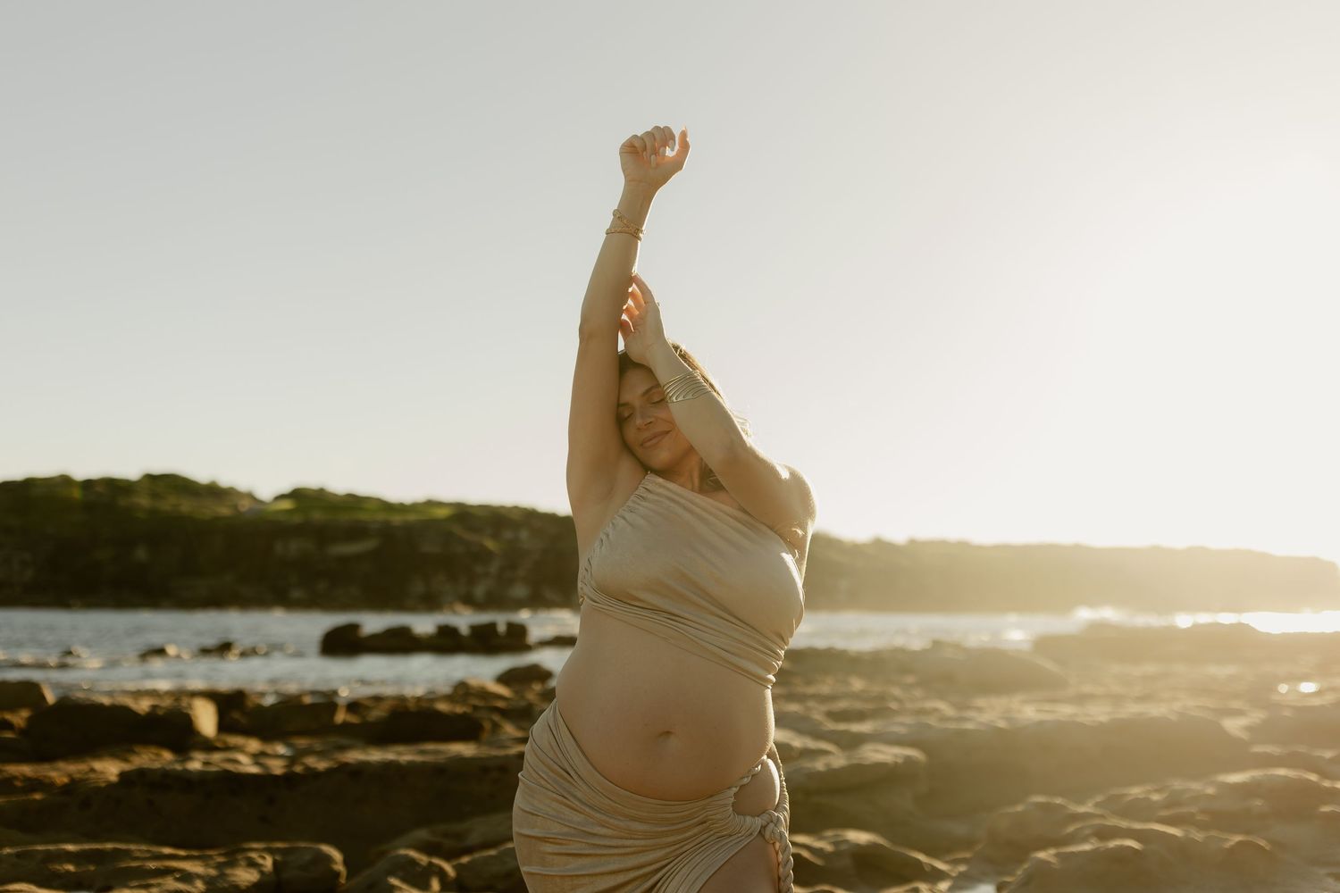 Pregnant woman in draped fabric poses on rocky beach at sunset with arms raised, glowing in warm golden light.