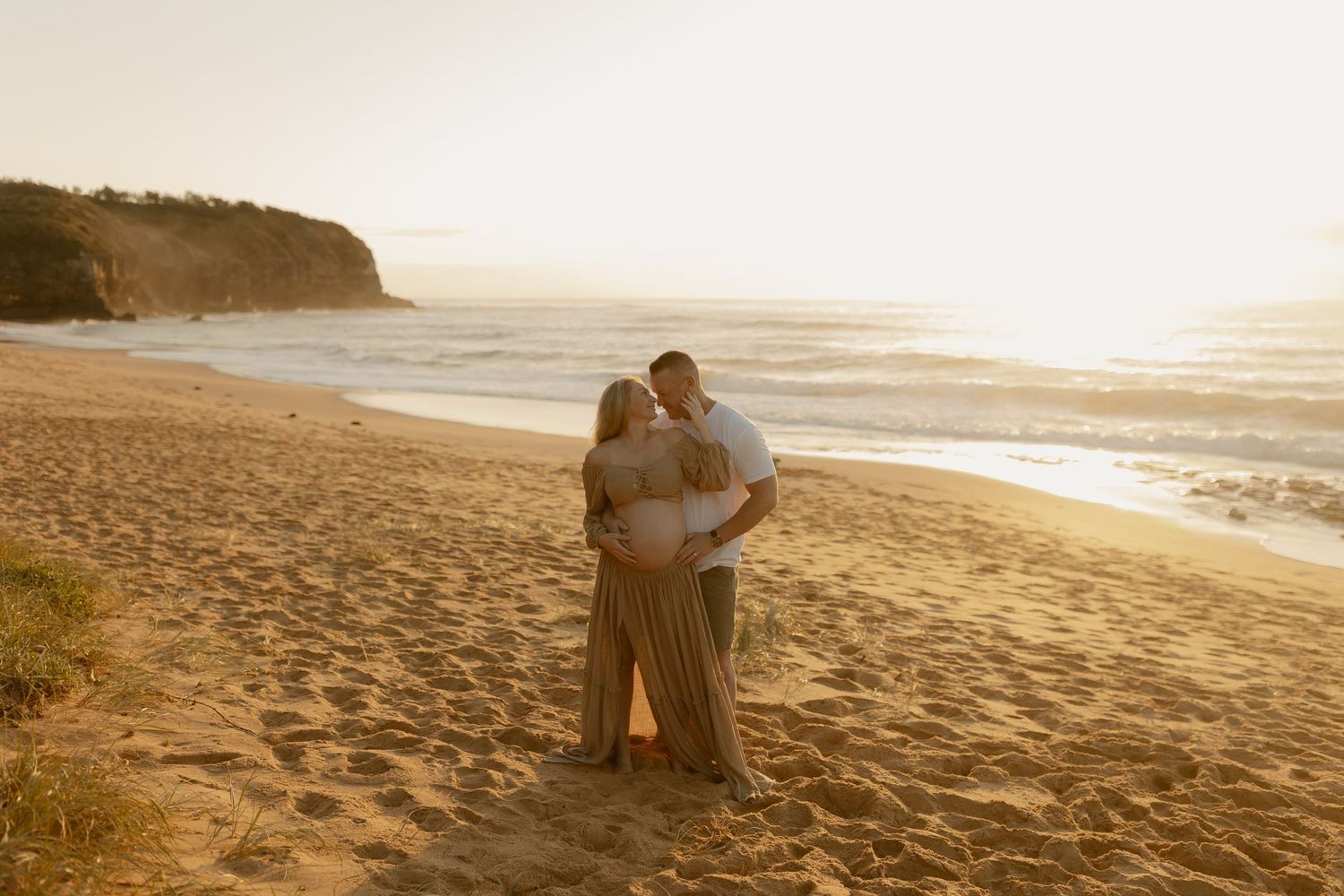 Couple embracing on a sunlit beach at golden hour, with ocean waves and sandy shores in the background.