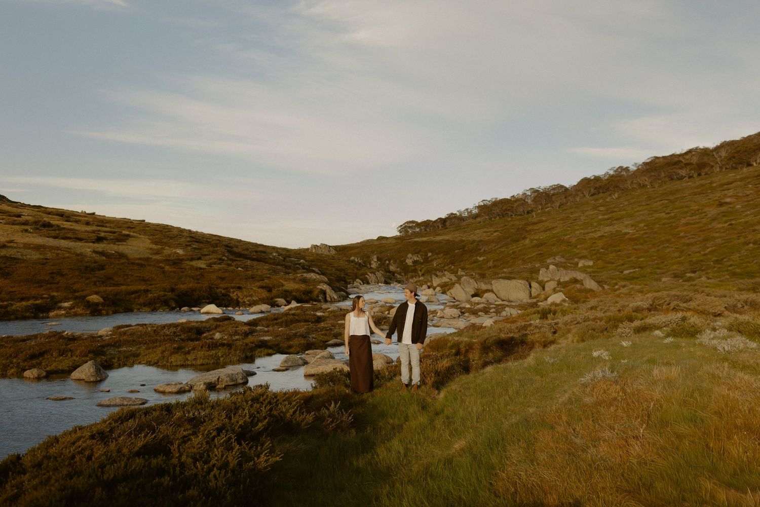 Couple in wedding attire embraces on a scenic hillside overlooking a coastal landscape at sunset.