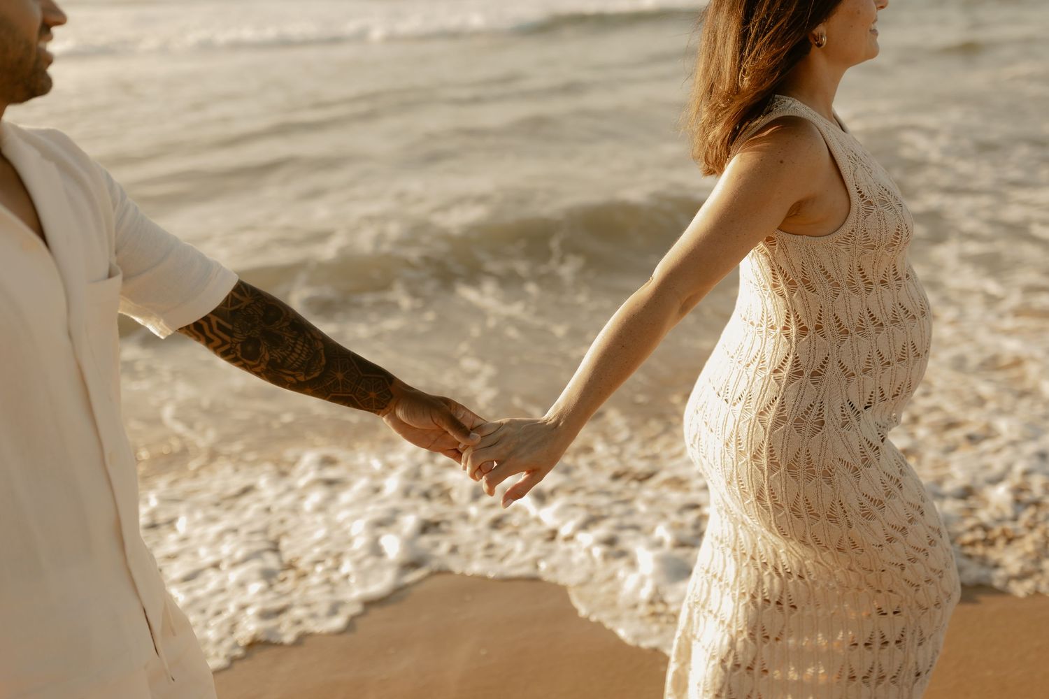 Couple holding hands walking along beach shore at sunset with woman wearing white lace dress.