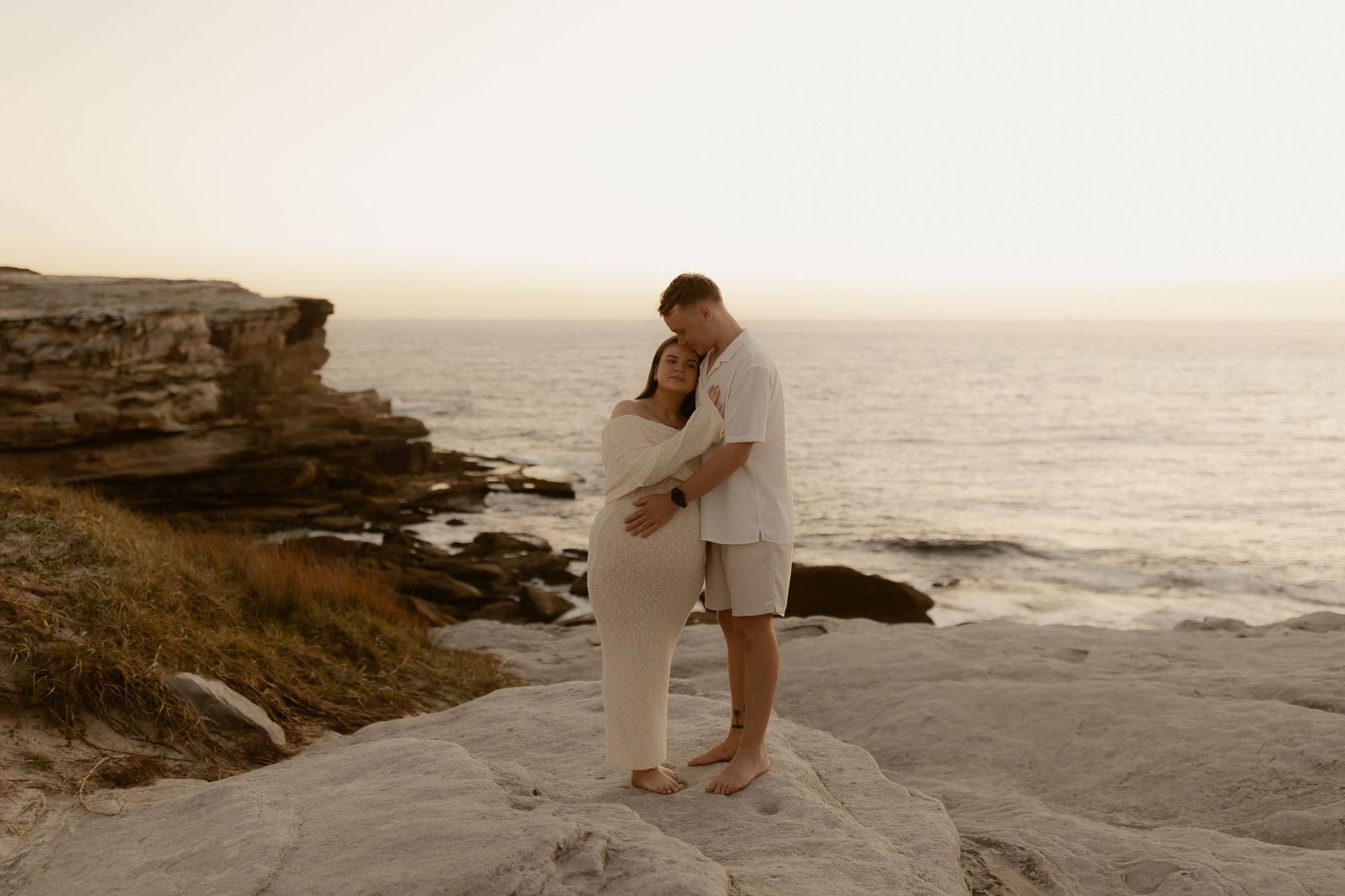 A couple embraces on a rocky coastal cliff at sunset overlooking the ocean.