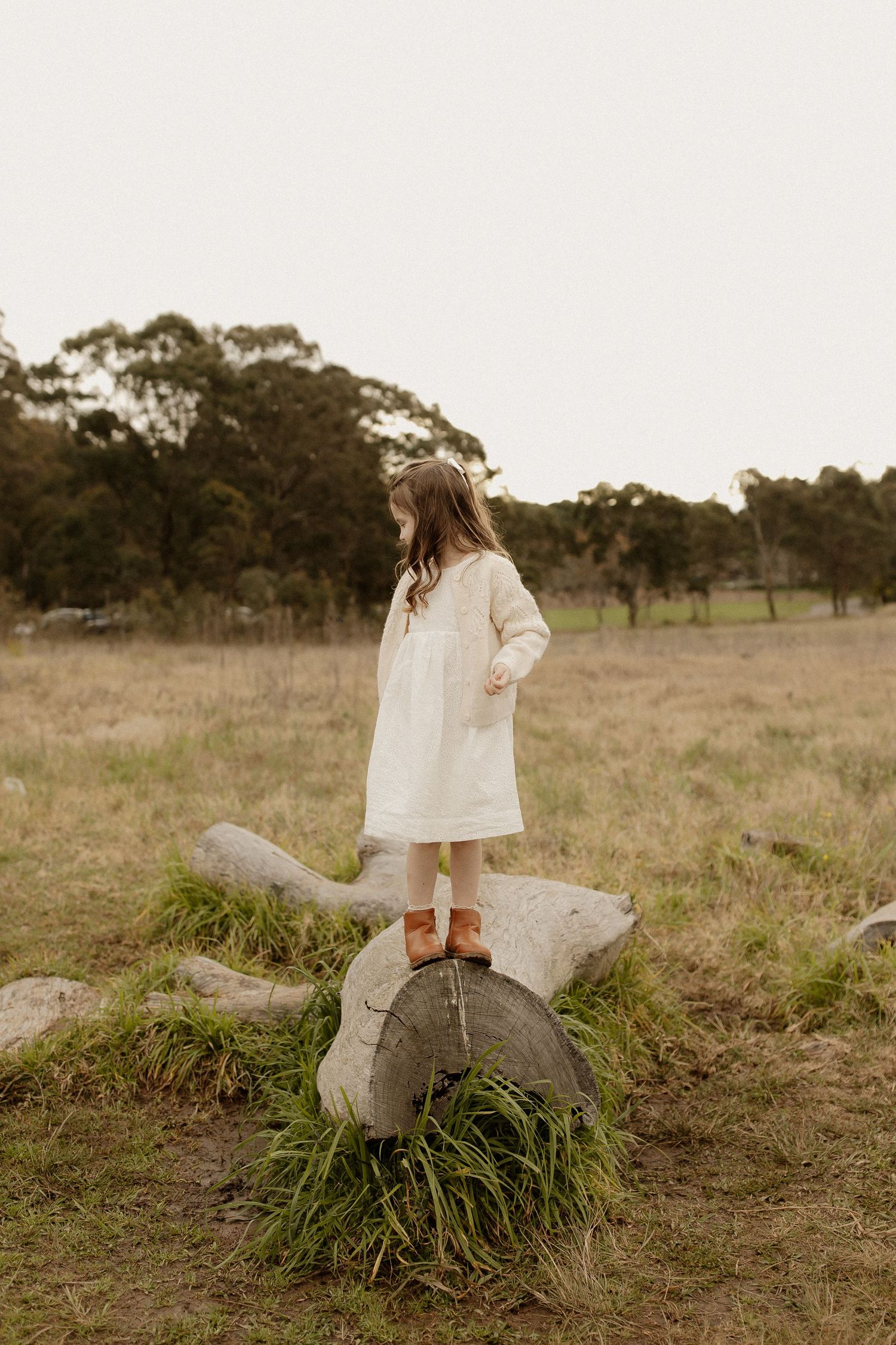 Person in flowing white dress sits serenely on a grassy knoll in a rustic outdoor setting.