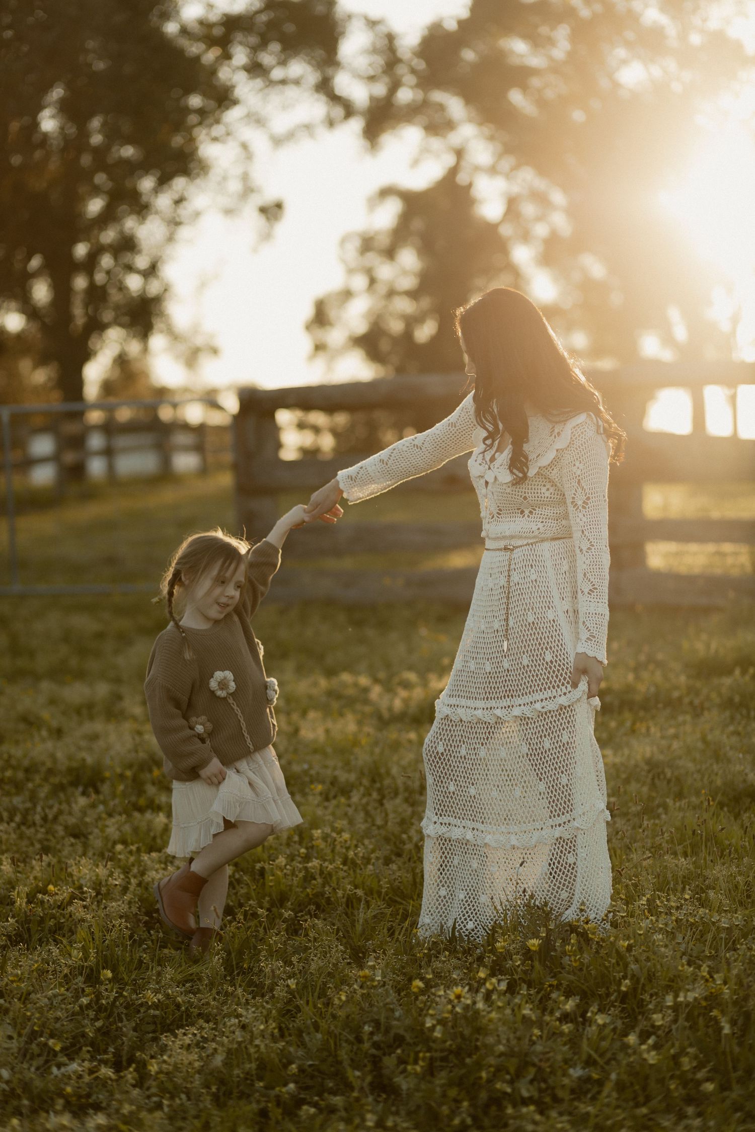 Woman in delicate white dress holds hands with small child in grassy field at sunset.