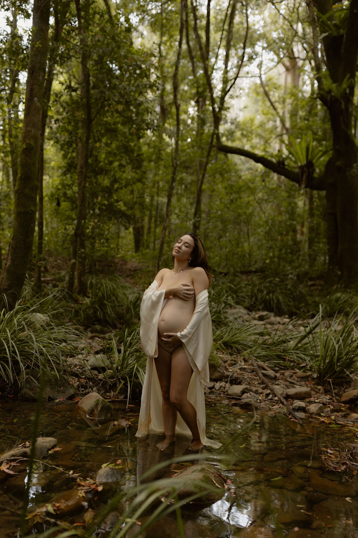 A dreamy portrait in a forest stream shows a figure in a beige cardigan posing thoughtfully among rocks and trees.