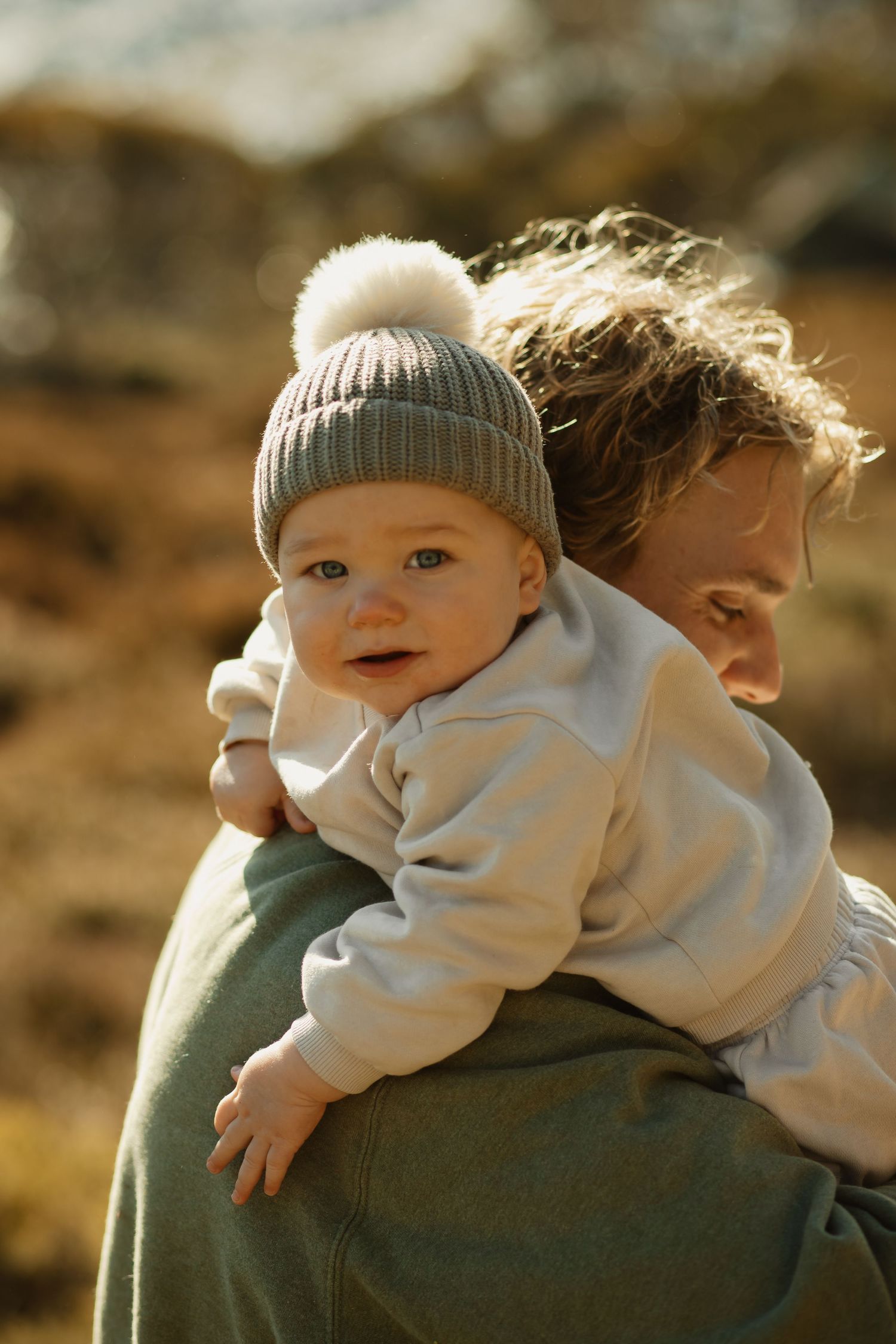 Baby wearing a knit pompom hat and white sweater being carried outdoors during golden hour light.