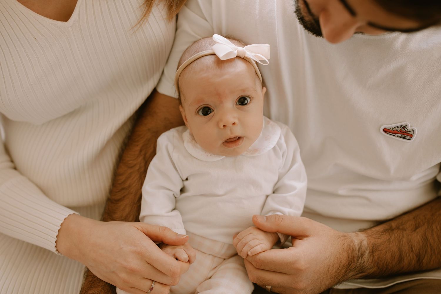 Small baby wearing white onesie and pink bow while being held between two people in white sweaters.