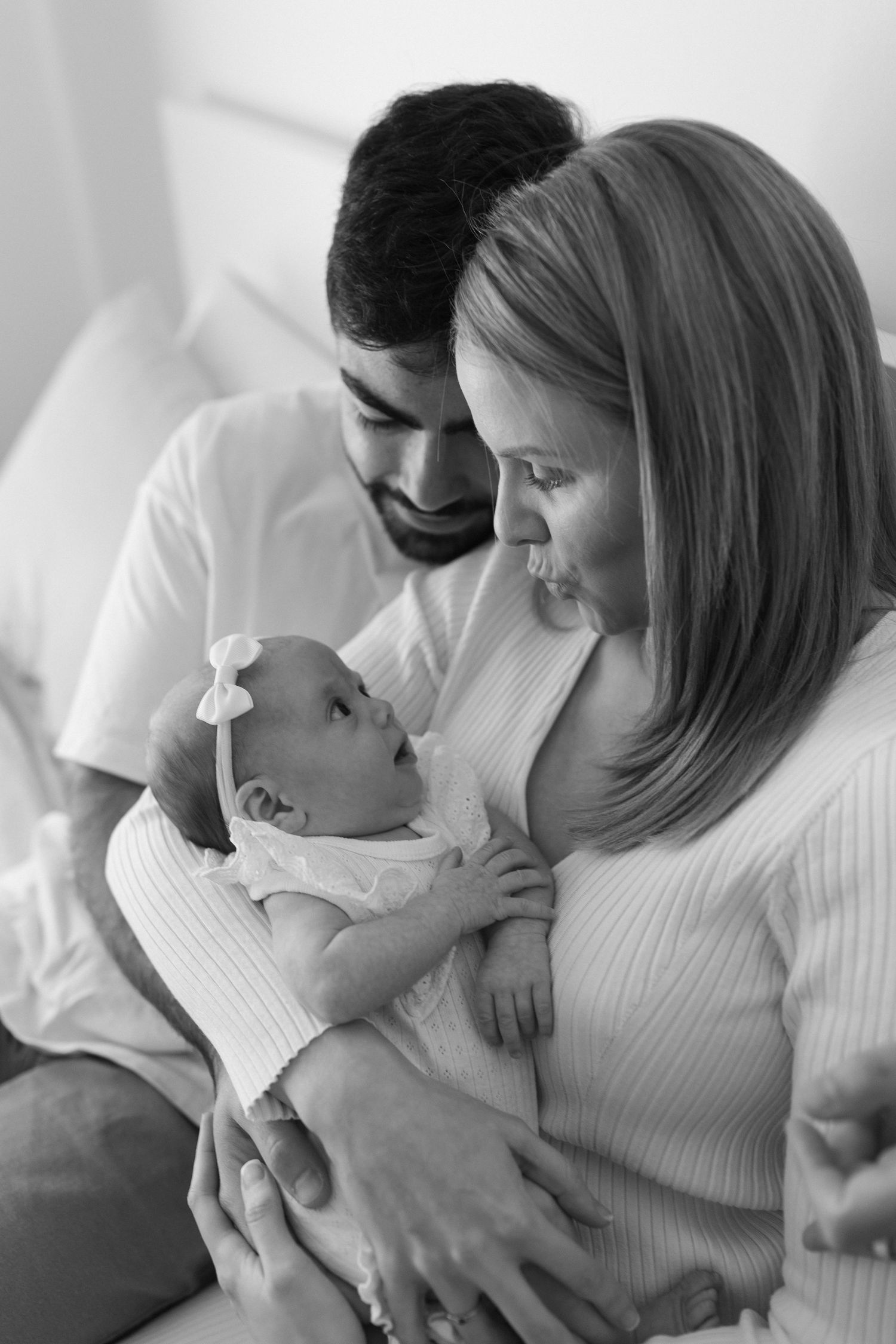 A black and white intimate photo of new parents gazing lovingly at their newborn baby wearing a bow.