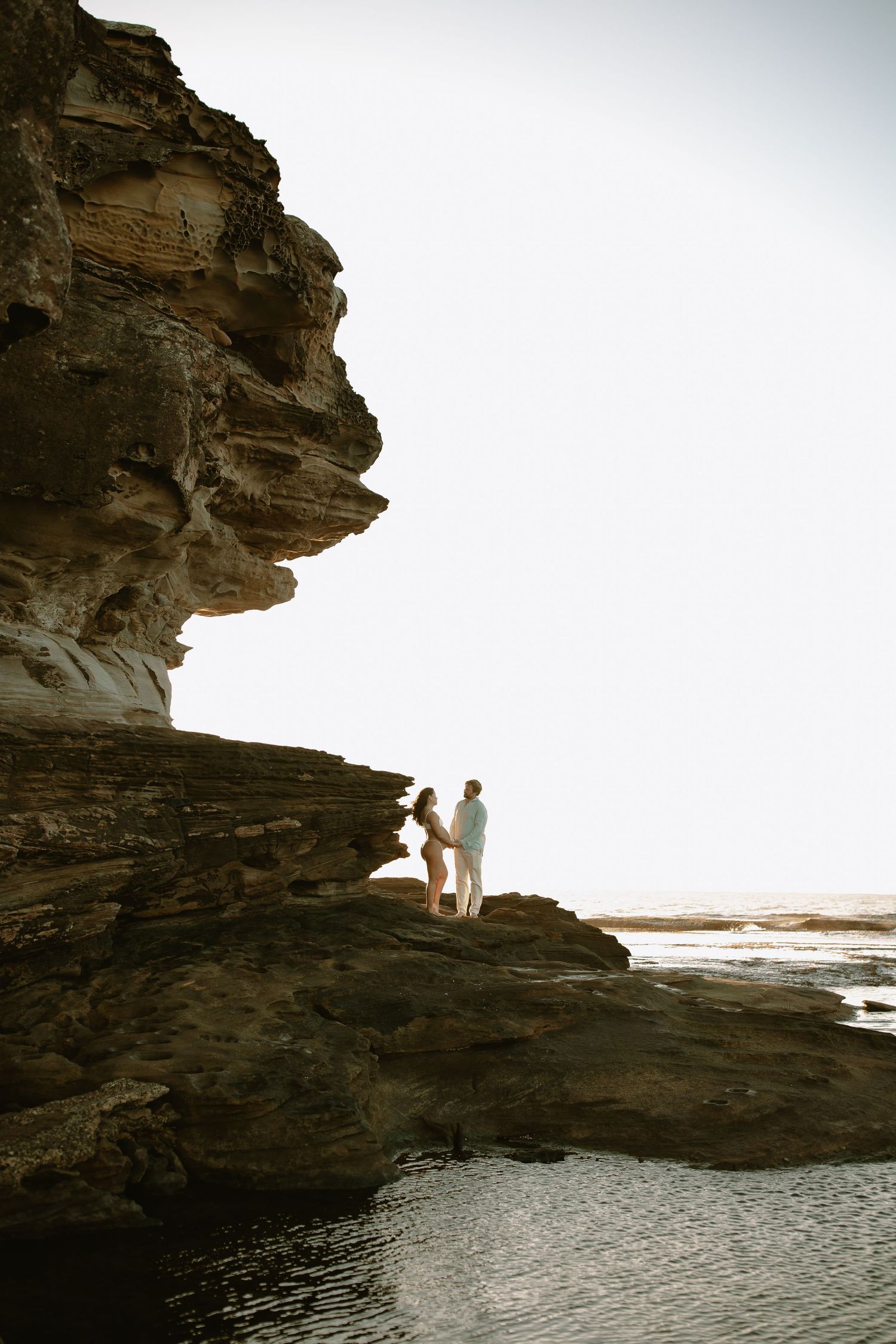 Two figures standing on a dramatic rocky cliff overlooking the ocean at sunset.