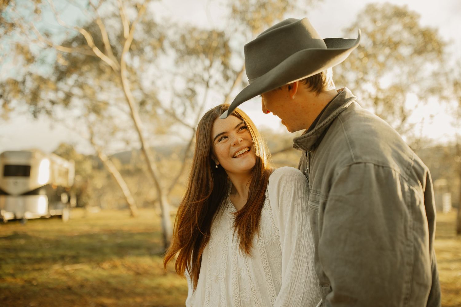 A couple shares an intimate moment in warm sunlight on a rustic campground with a trailer in the background.