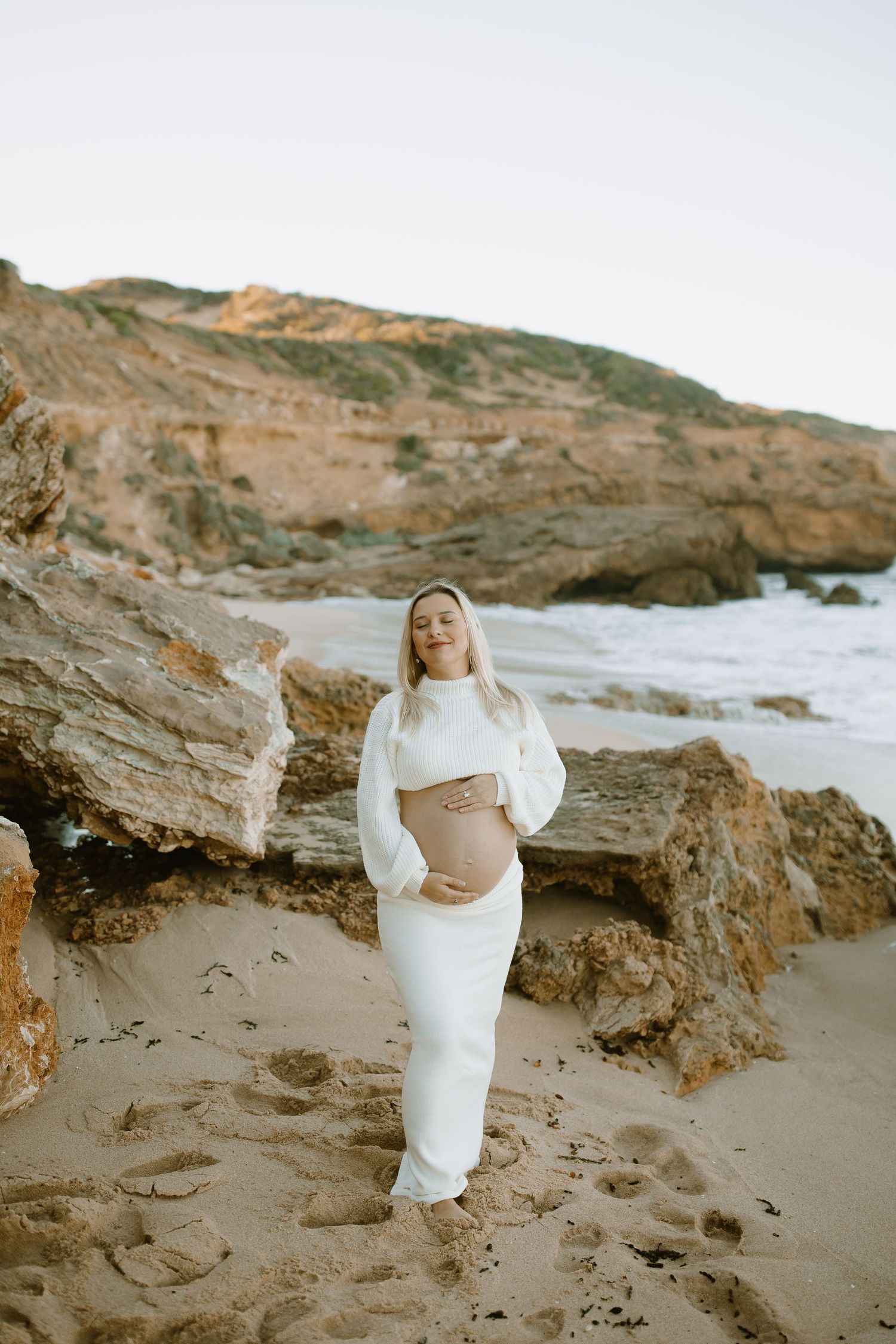 A maternity photoshoot on a rocky beach with cliffs and ocean waves in the background.