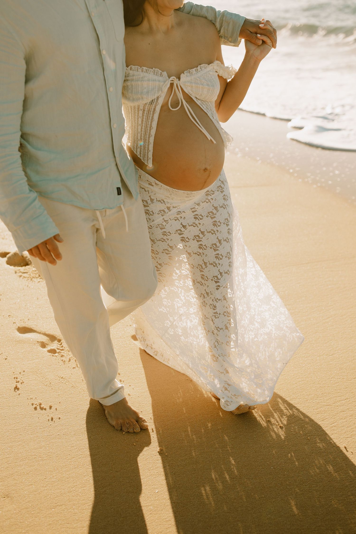 Expecting couple in white lace maternity dress and light colored suit standing on beach sand at golden hour.