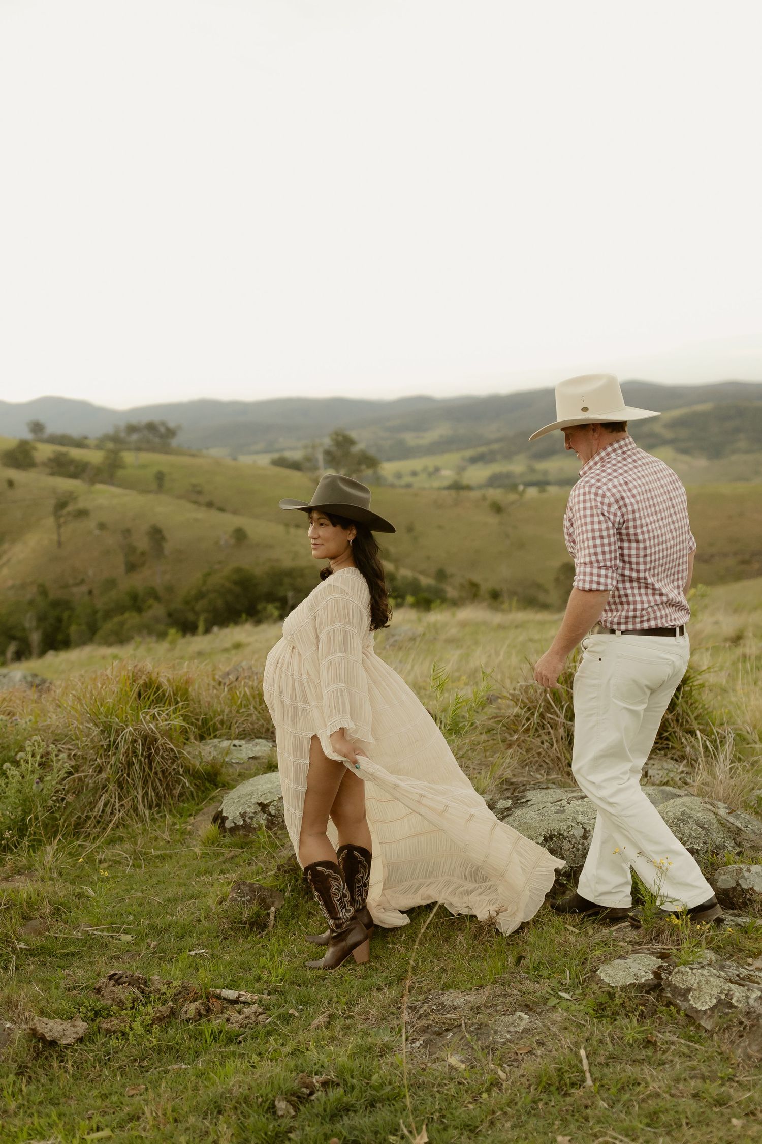 A couple in cowboy hats hold hands on a grassy hillside, woman in white dress and boots, man in plaid shirt.