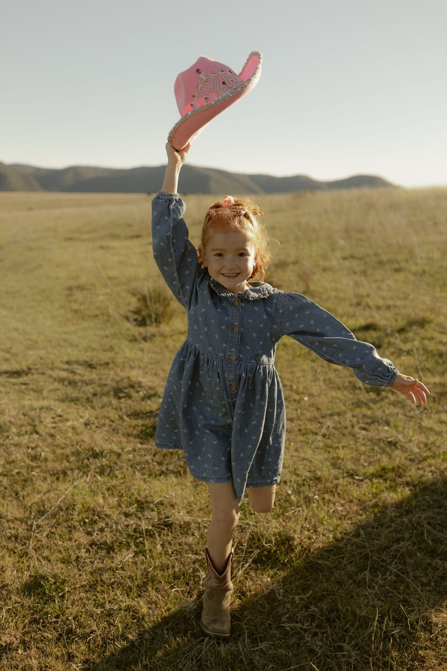 A joyful little girl in a denim dress raises a pink cowboy hat while playing in a sunny green field.