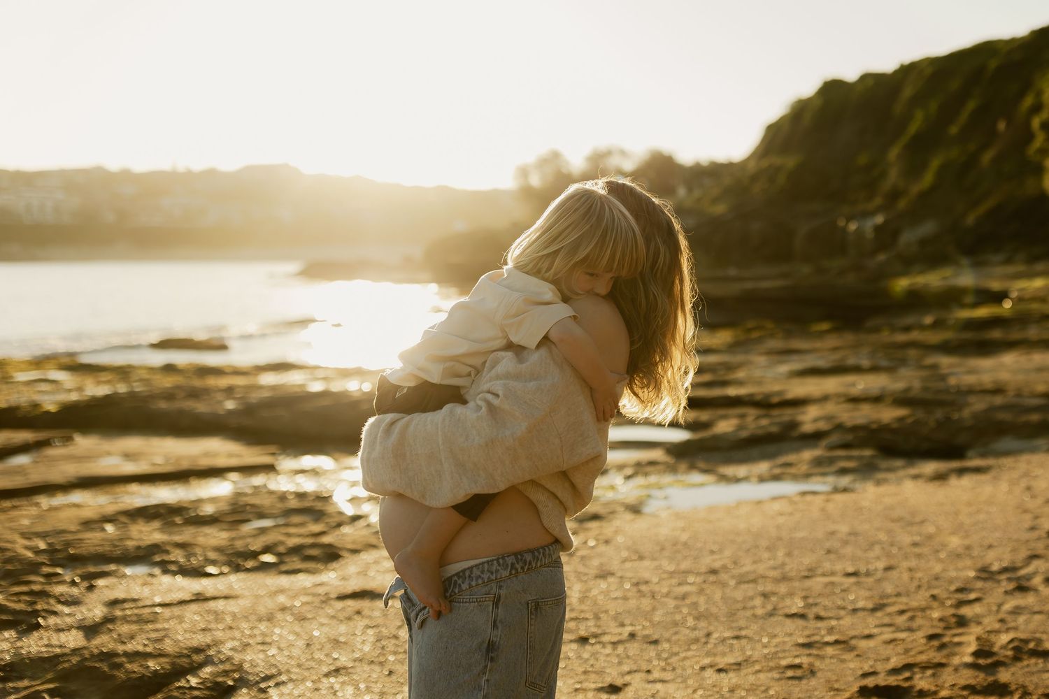Mother tenderly hugging young child on sandy beach during warm golden hour sunset light.