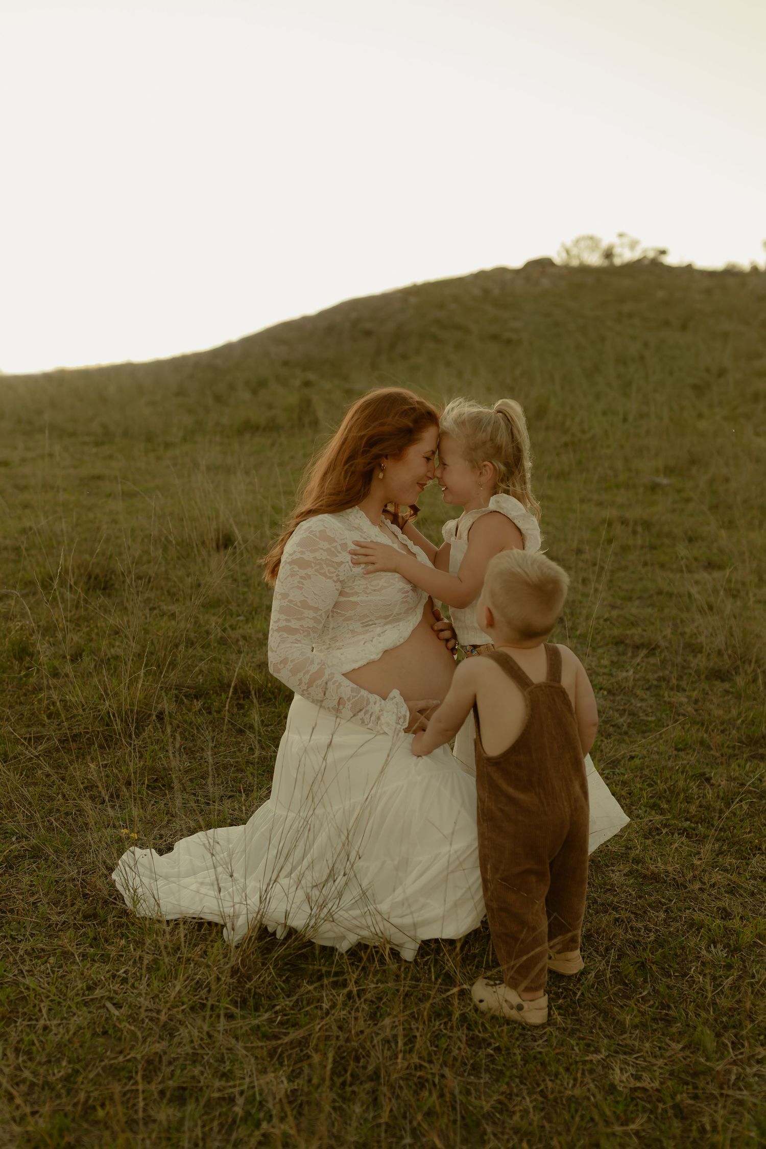 Pregnant woman in white lace dress kneeling on green hillside, sharing tender moment with two young children at golden hour.