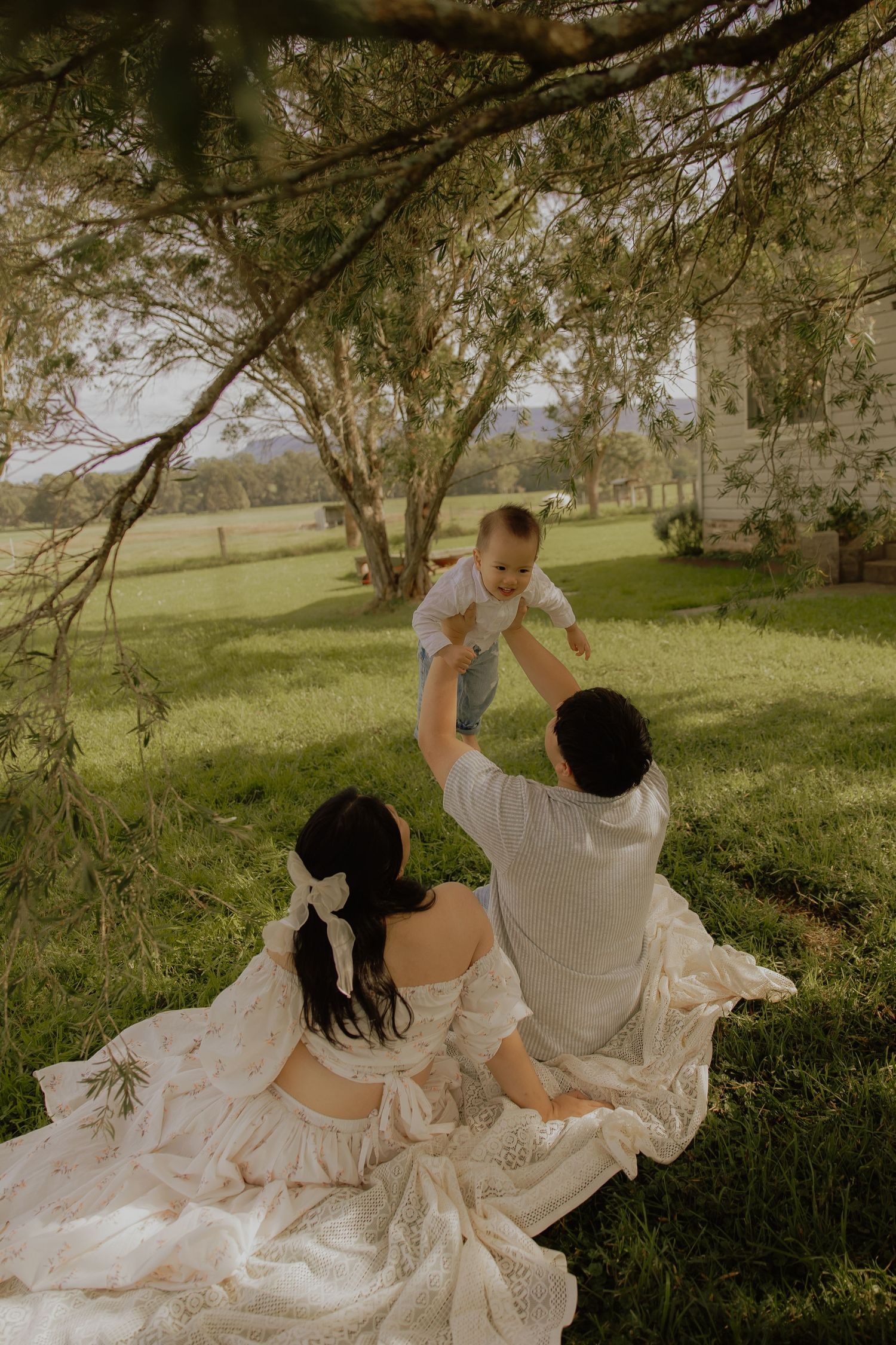 Parents sitting on grass lifting baby in air under tree, elegant outdoor family photo session in green meadow.
