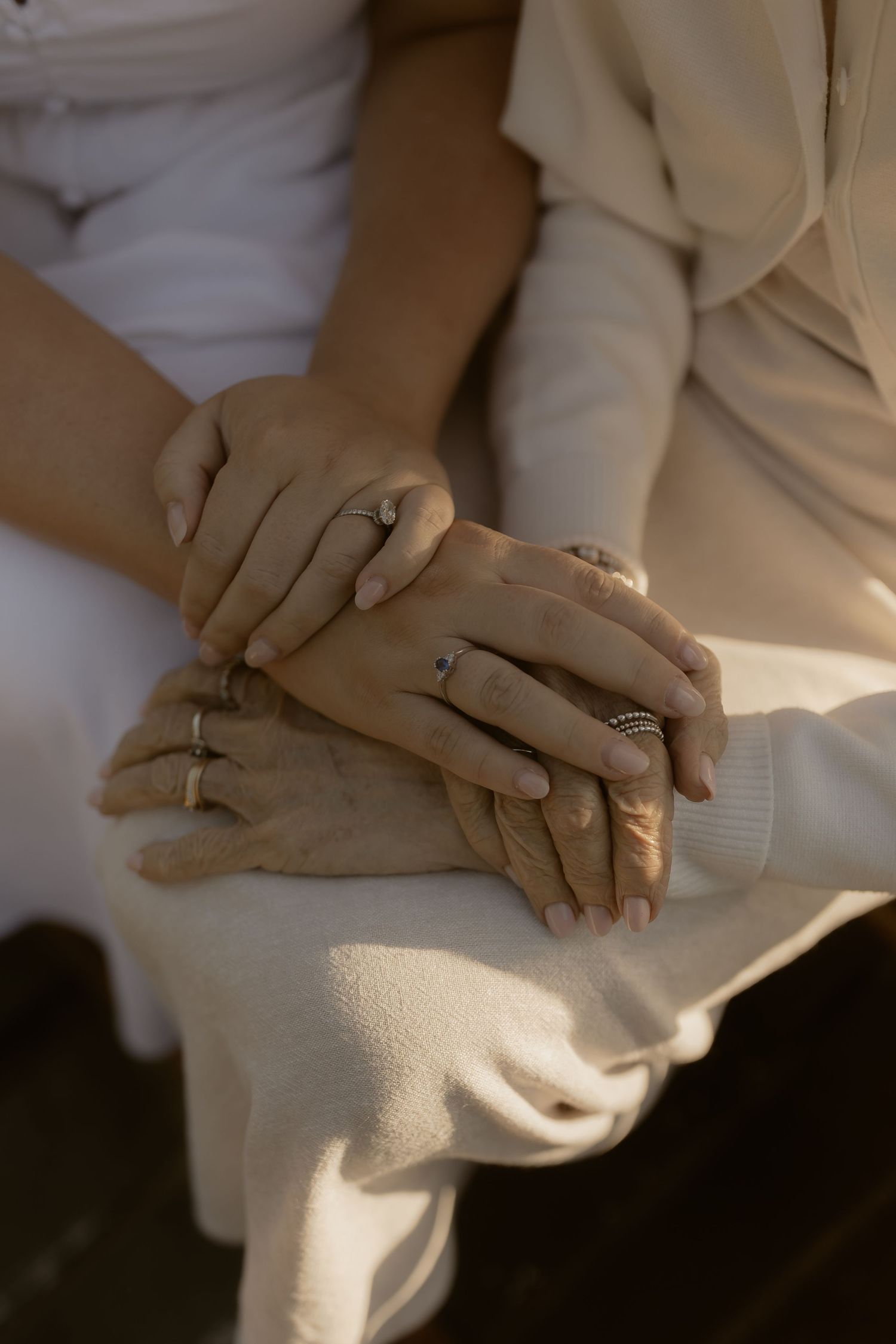 Close-up of two people holding hands with rings, wearing white and cream clothing in warm sunlight.