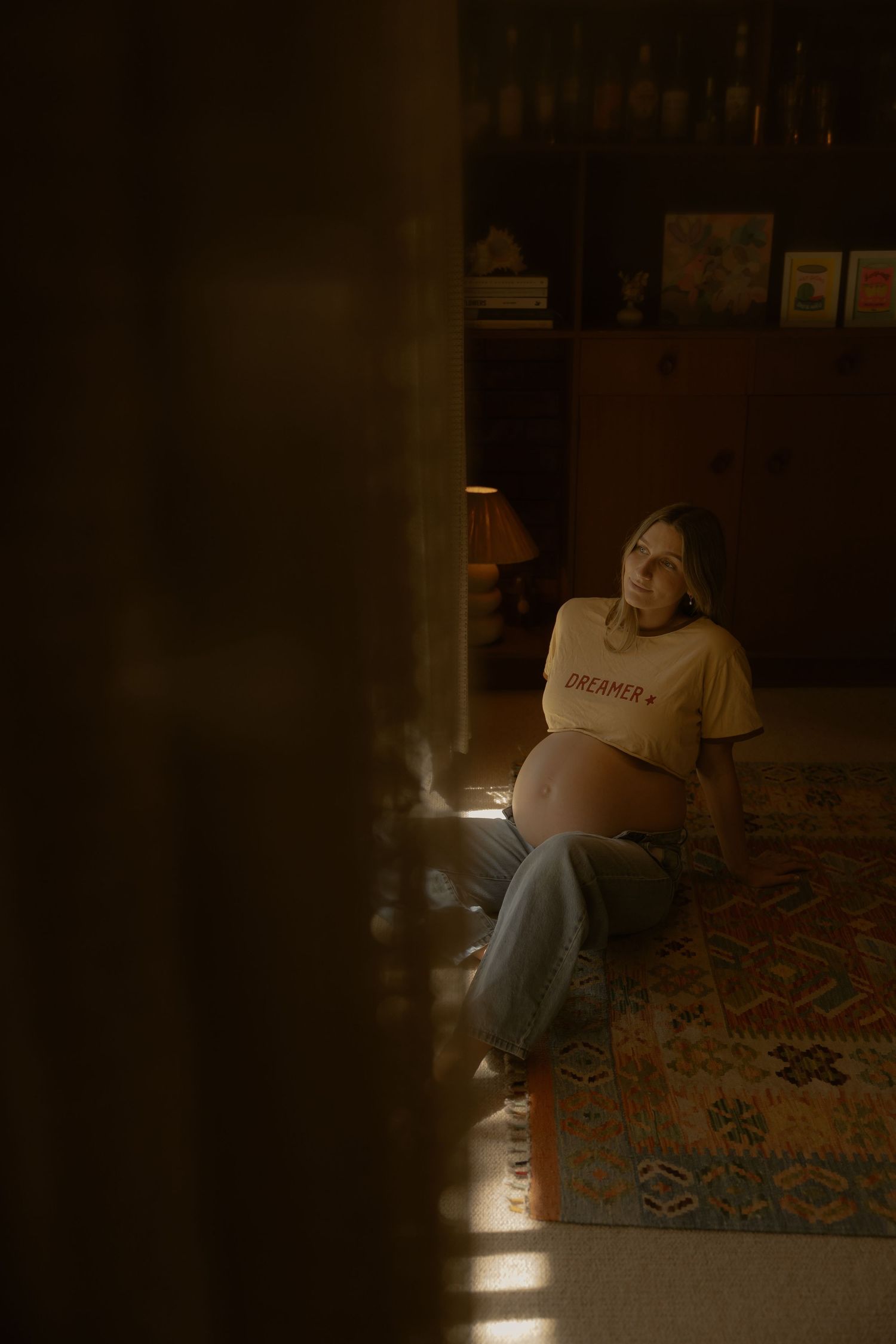 Pregnant woman in yellow Dreamer tee sits on patterned rug in warm moody light.