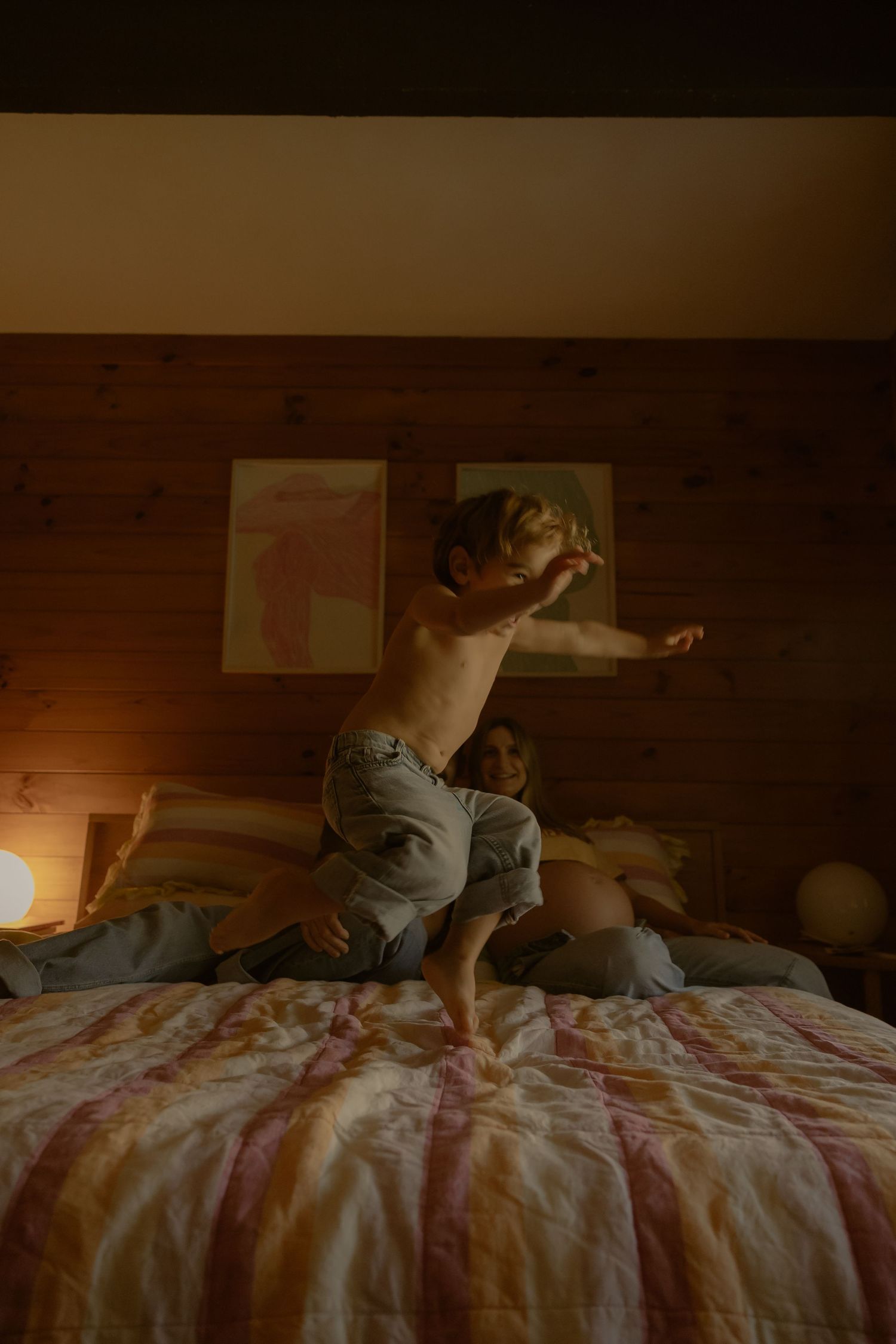 A shirtless toddler joyfully jumps on a bed in a cozy, warmly lit wooden bedroom.