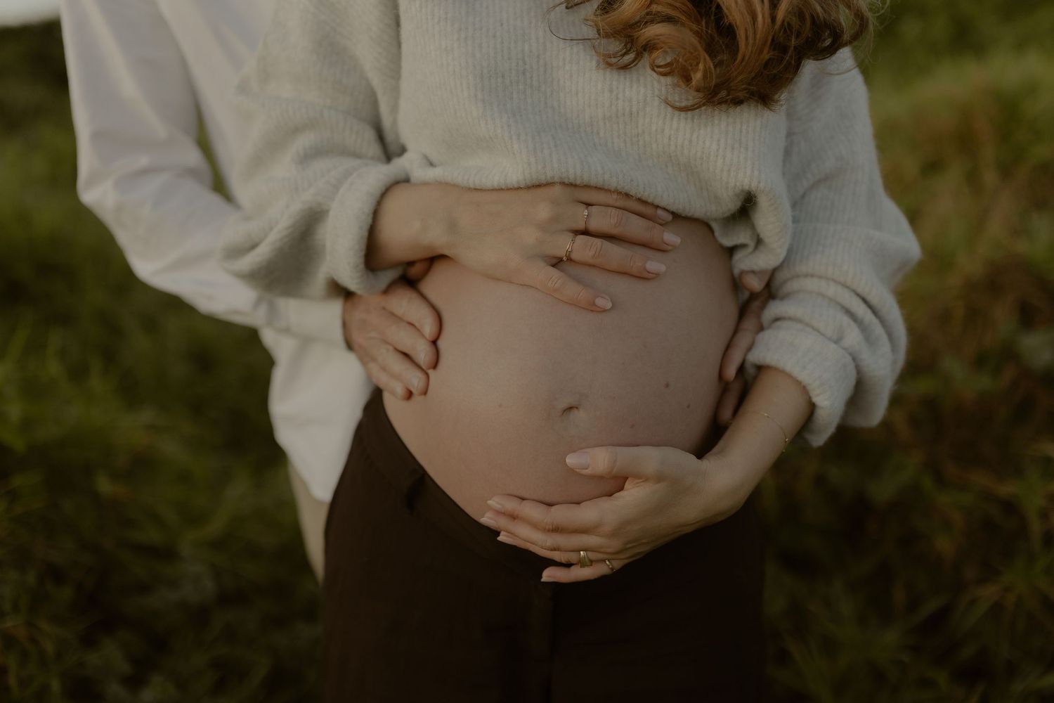 Close-up of pregnant woman's bare belly being cradled by two pairs of hands outdoors in green field.