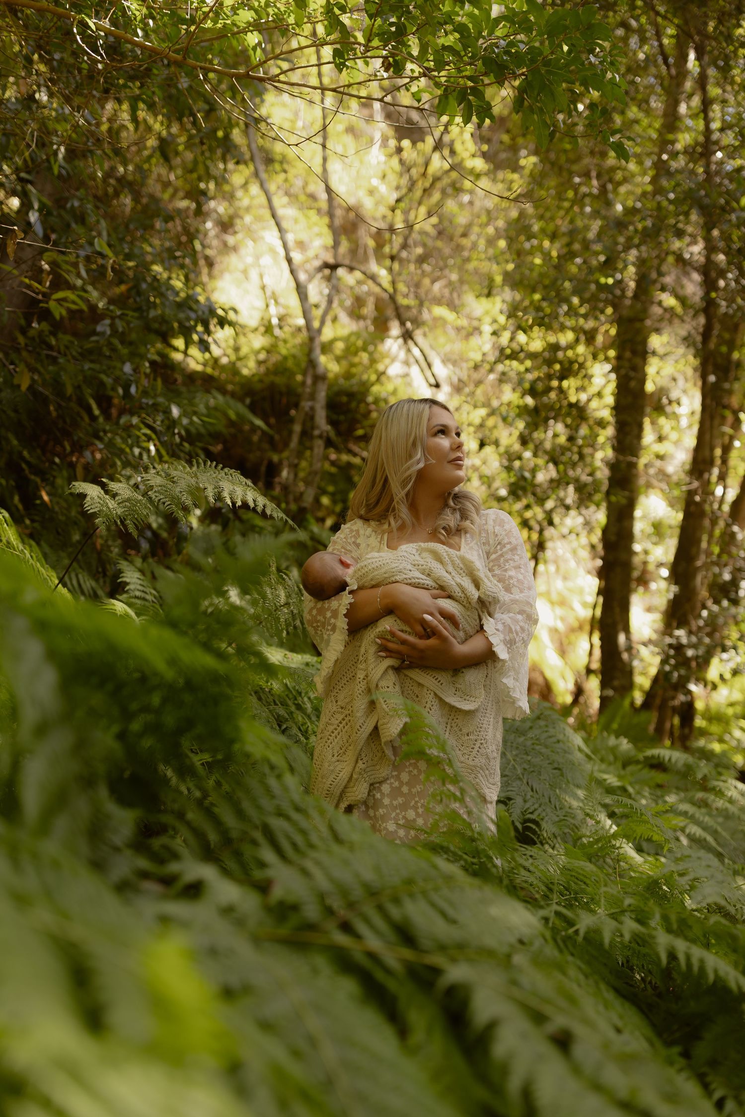 Blonde woman holding newborn baby in lush green forest surrounded by ferns, wearing white lace dress.