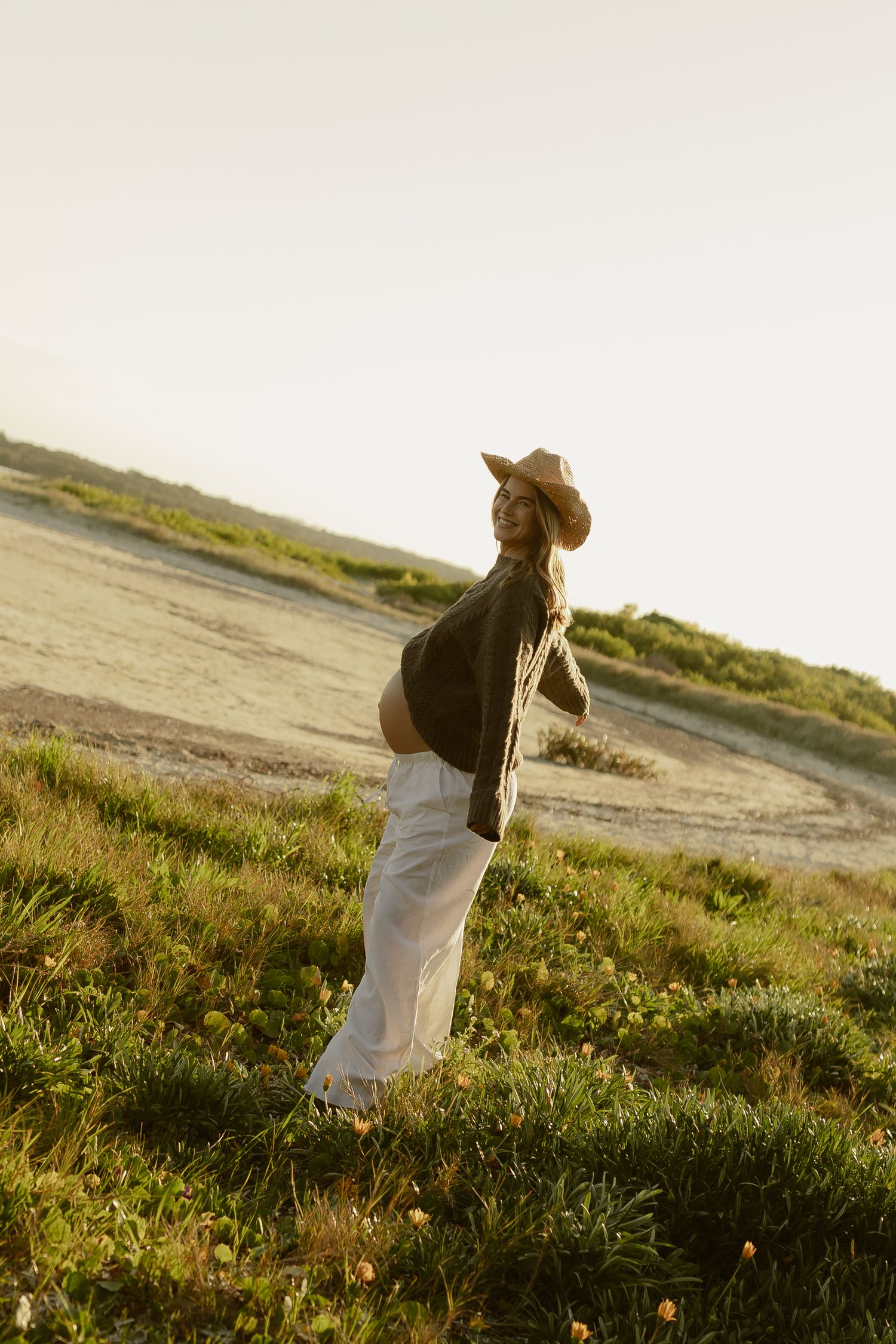 Pregnant woman in cowboy hat, dark top, and white pants standing in a sunlit grassy field during golden hour.