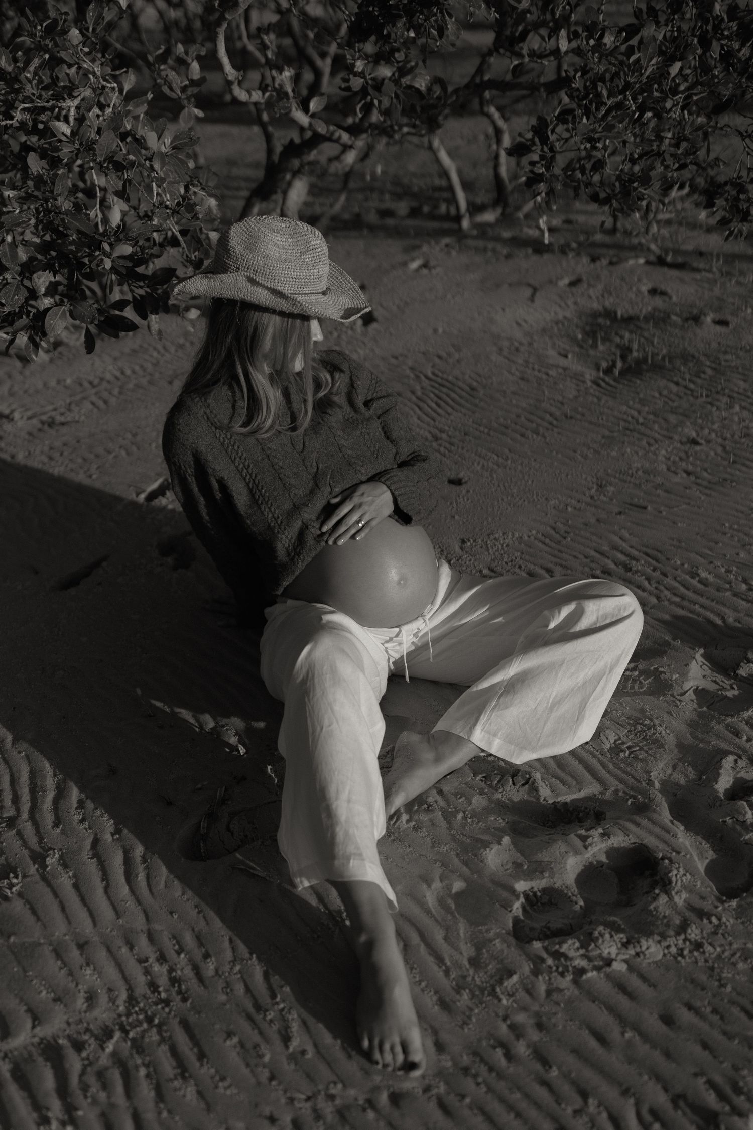 Black and white photo of a pregnant woman sitting on sand, wearing a hat and white pants, cradling her bare belly.