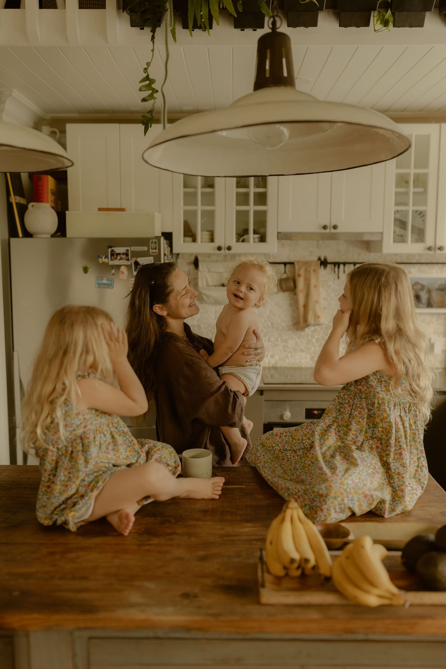 A mother holds her baby while two blonde girls in floral dresses sit on a wooden kitchen counter.
