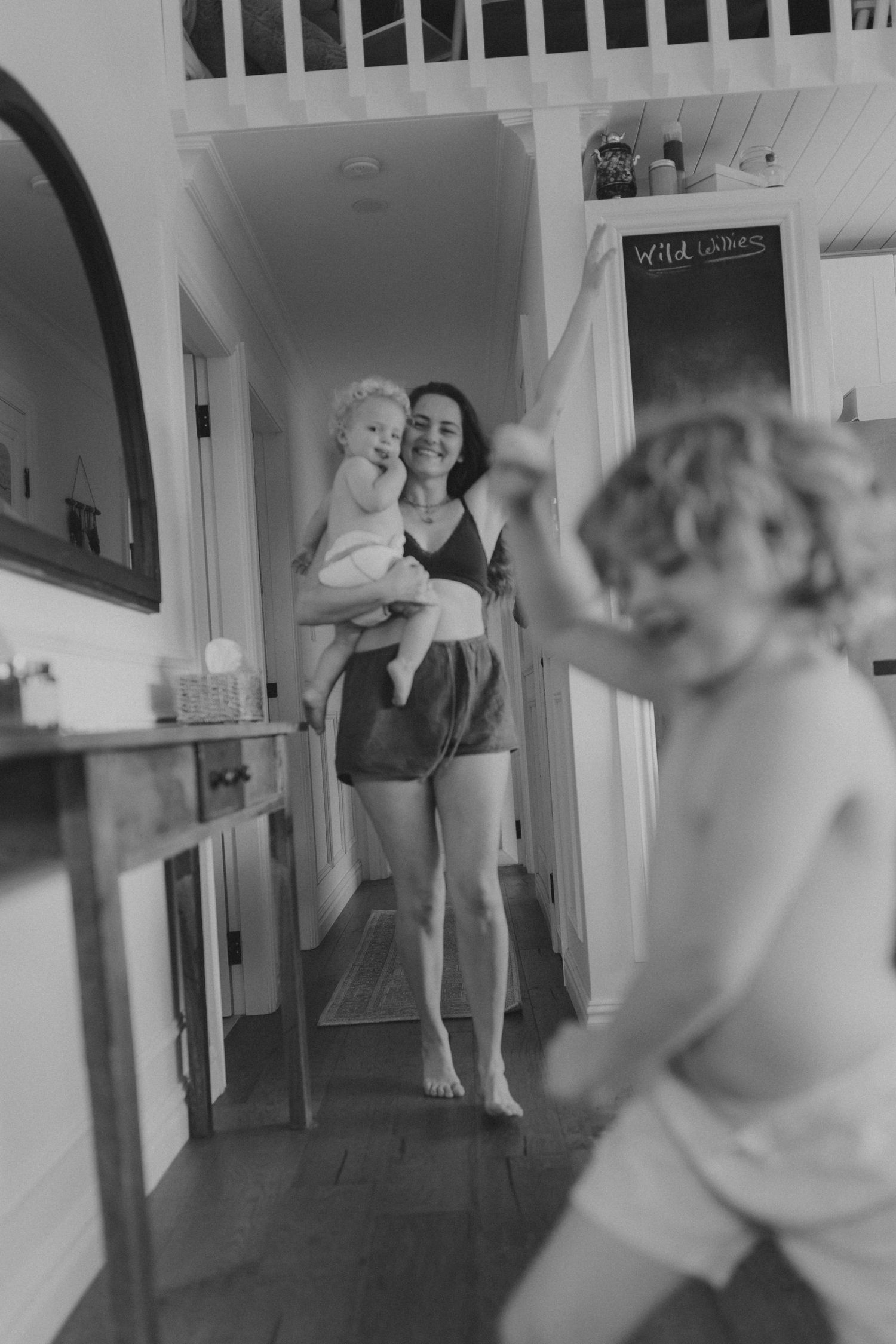 Black and white photo of a smiling mother holding a baby while a toddler plays in a bright hallway.