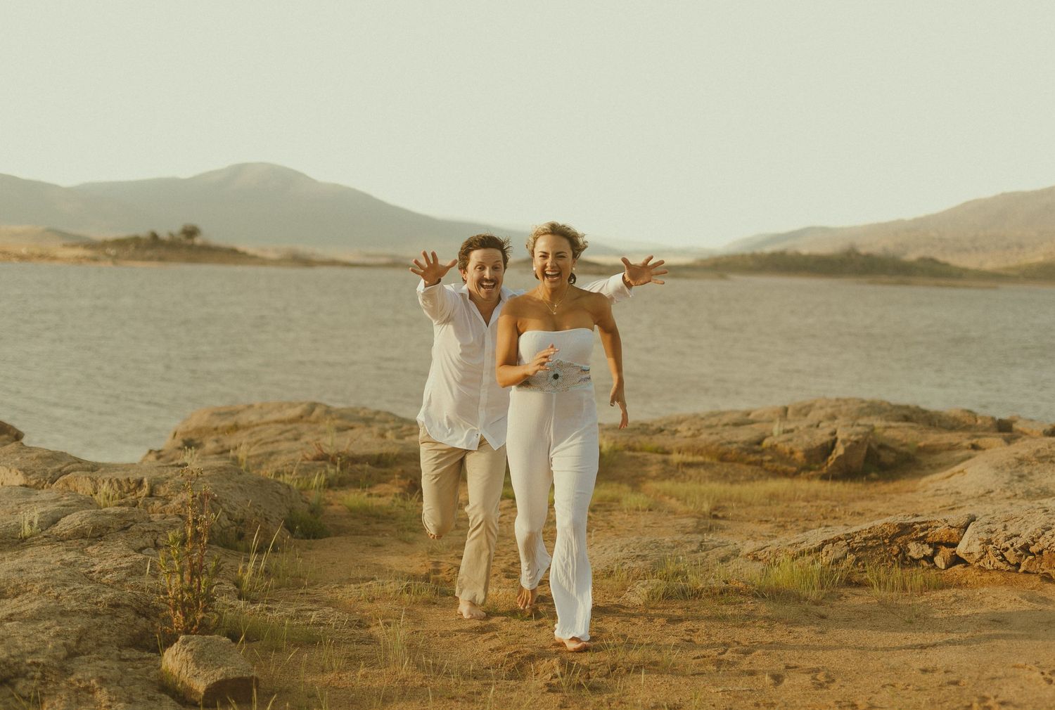 Two people in white outfits pose joyfully on rocky terrain by a lake with mountains in the background at sunset.