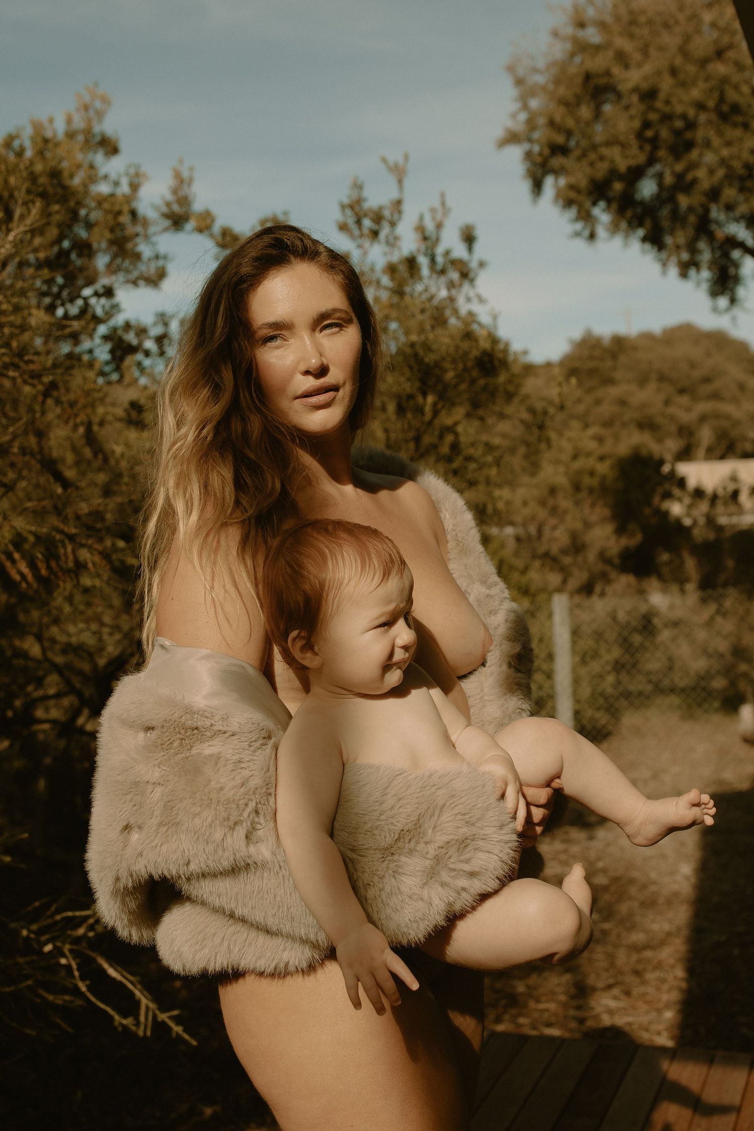 A mother in beige clothing holds her baby outside during golden hour with trees in the background.