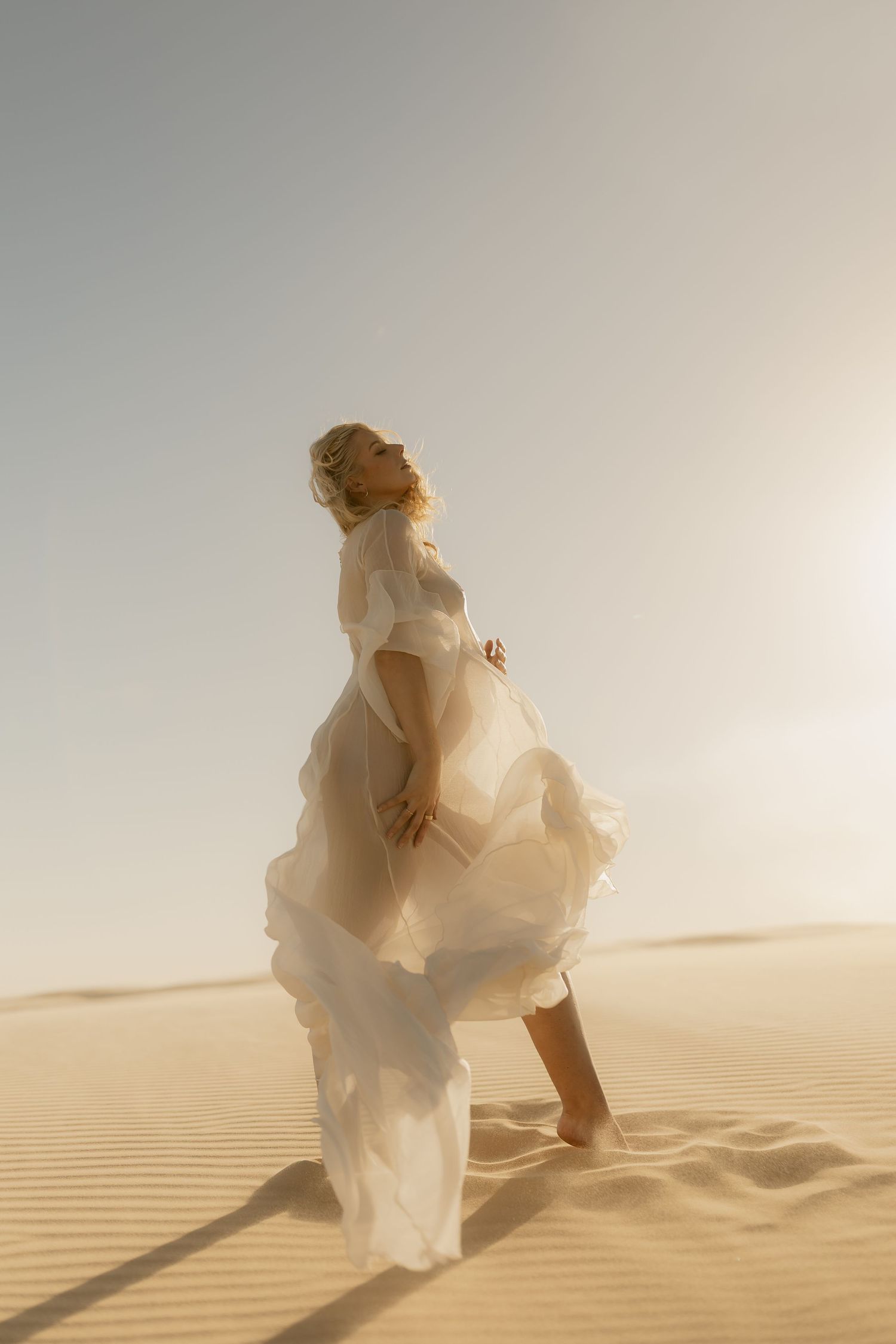 Pregnant woman in flowing white dress standing on golden sand dunes in desert sunlight.