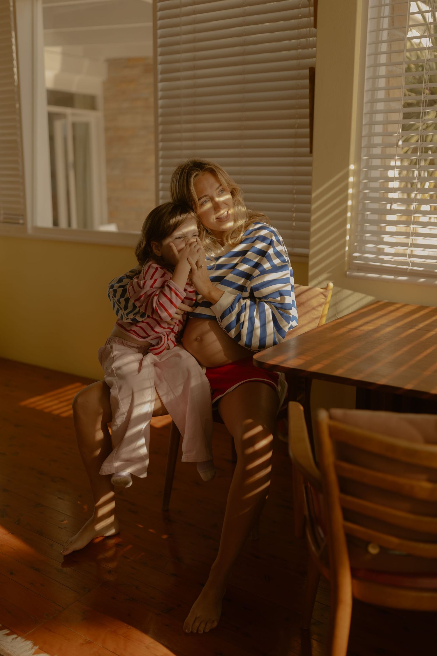 Mother and child sitting together at wooden dining table in warm sunlit room with striped blinds casting shadows.