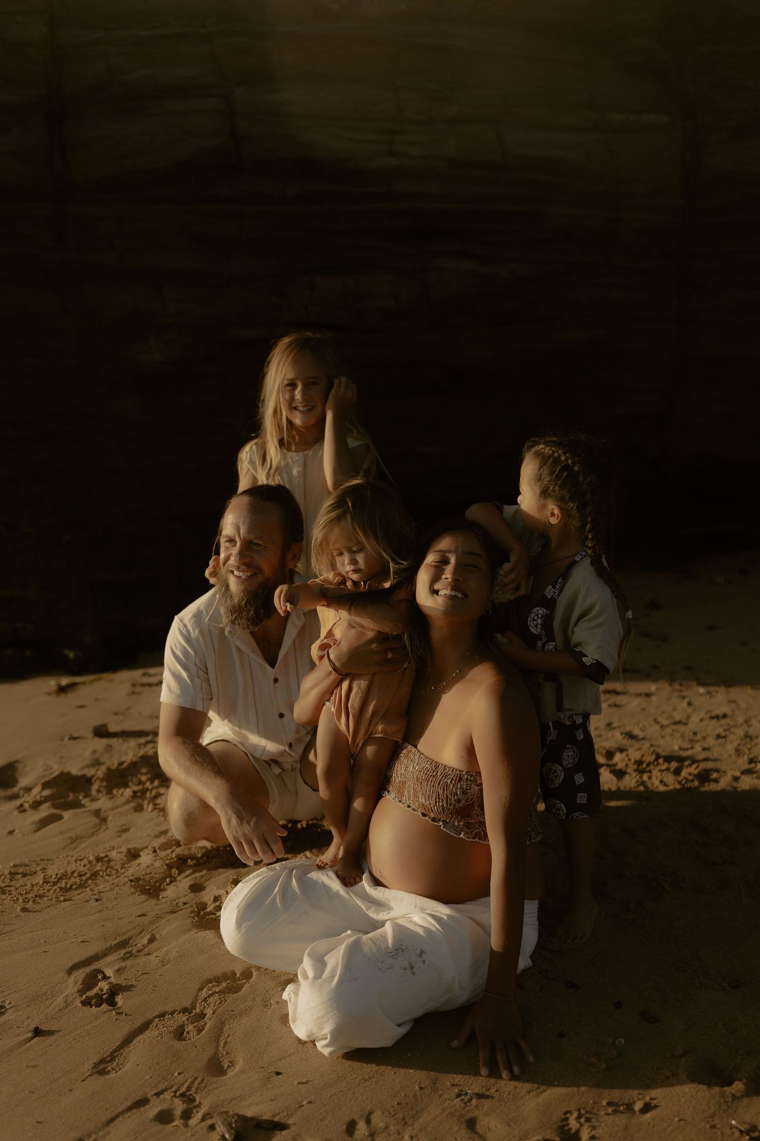 A happy family with three young children sits together on sandy beach near a dark cave during golden hour.