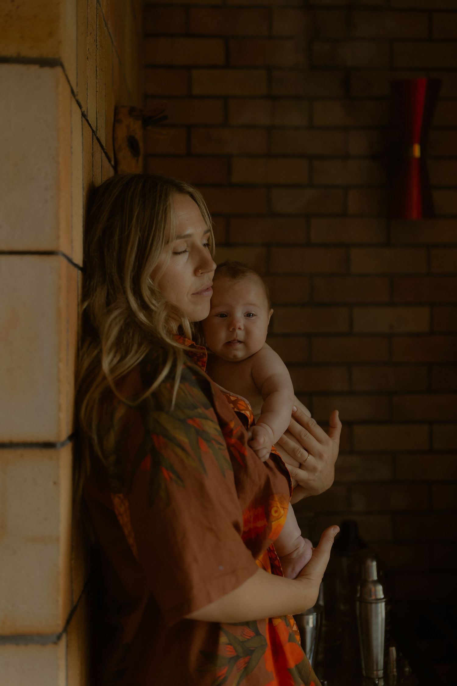 A mother in an orange floral shirt tenderly holds her newborn baby against a rustic brick wall indoors.