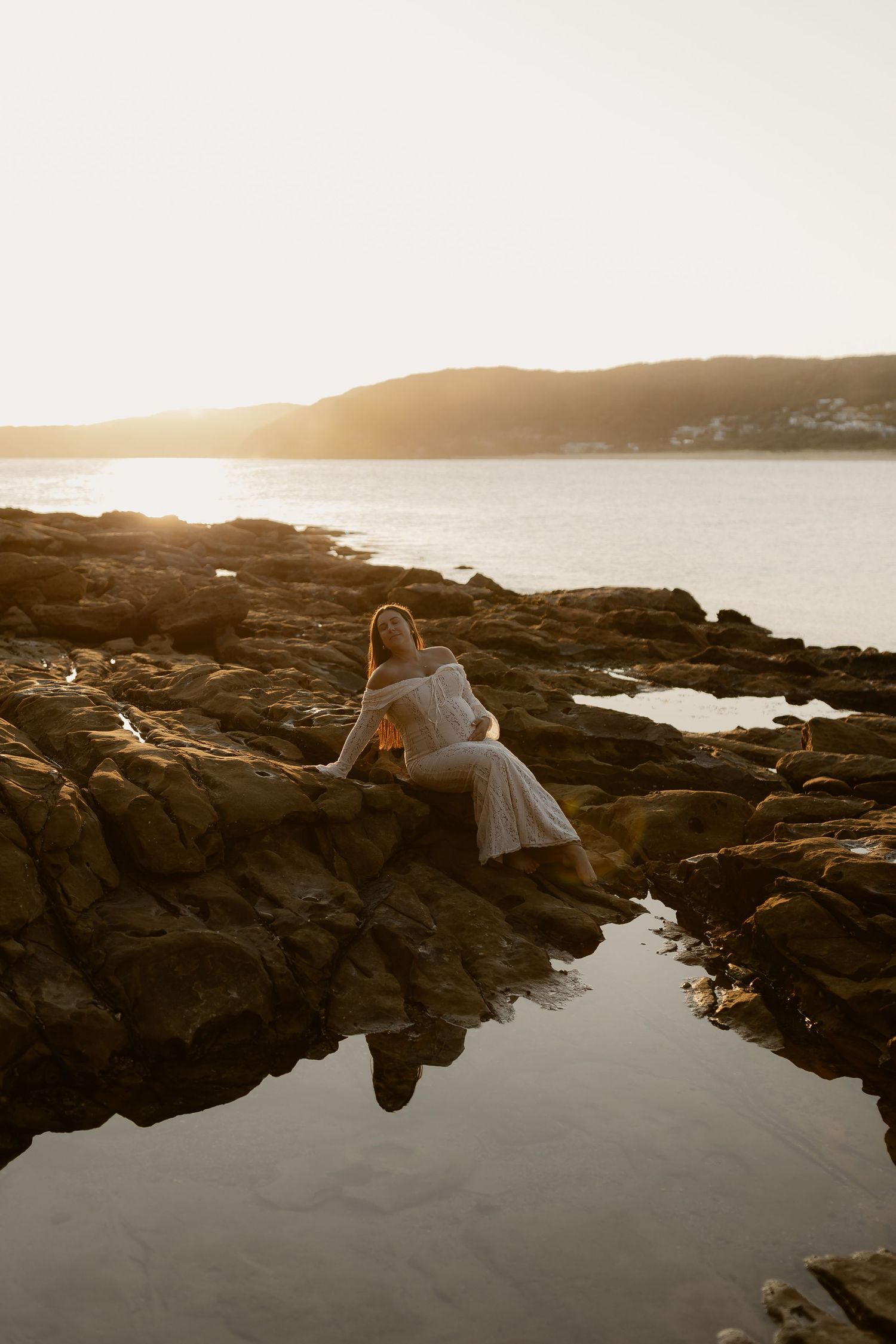 Woman in white dress sitting on rocky shore at golden sunset with calm ocean and hills in background.